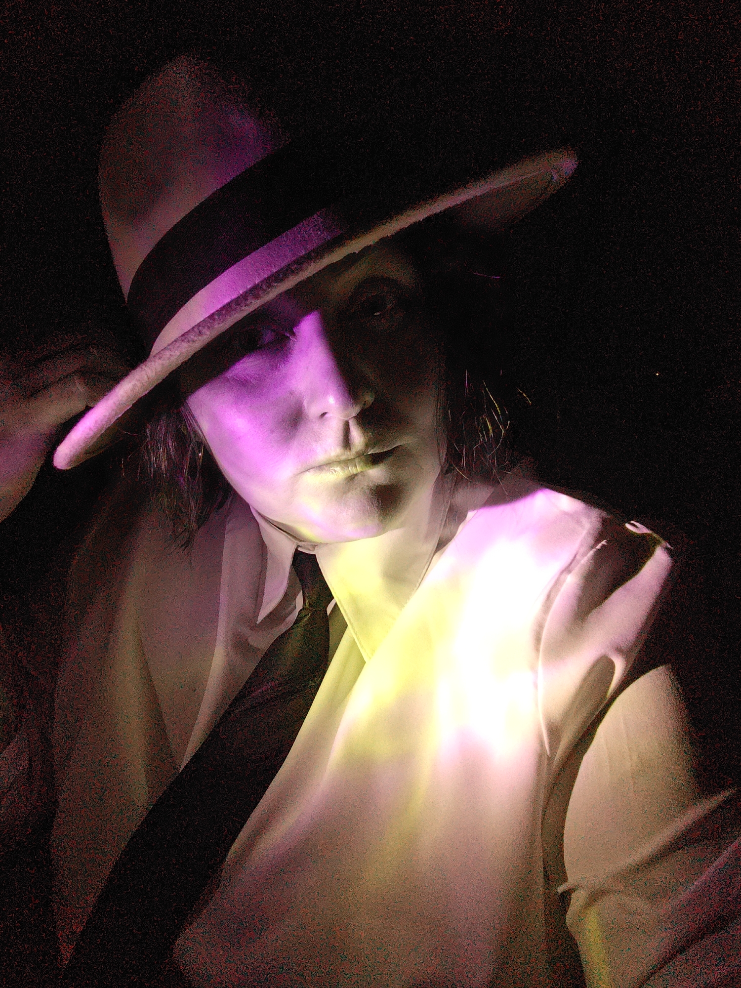 A darkly lit, portrait-style photograph features an individual wearing a fedora-style hat, a white collared shirt, and a dark tie. The person is shown from the chest up, looking directly forward with a serious and direct expression. Their left hand is subtly touching the brim of the hat.
The subject's face is dramatically illuminated by a magenta-purple light source coming from the left, highlighting the cheekbone, nose, and lips, while much of the right side of the face and the underside of the hat brim are cast in deep shadow. A separate, bright yellow-green light source illuminates the individual's right shoulder and chest, creating a striking color contrast with the purple light on the face.
The background is uniformly dark and undefined, drawing all focus to the subject and the interplay of light and shadow. The image possesses a noticeable grainy or noisy texture, especially in the darker areas. No text is visible. The scene suggests a posed shot, likely taken indoors under controlled, artificial lighting, rather than in an outdoor or naturally lit environment. There are no specific visual cues within the image that point to the provided location context of Markovskii, Russia.