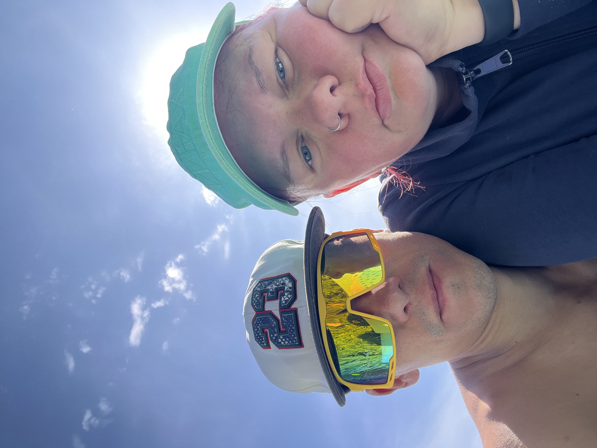 Two individuals are in the frame, shot from a low angle against a backdrop of a blue sky with scattered clouds and the sun. The person at the top of the frame, with fair skin, is wearing a light green cap and a dark blue hoodie, with a nose ring visible. The person at the bottom of the frame is wearing a light-colored baseball cap with the number "23" on it and yellow sunglasses with reflective lenses. They appear to be shirtless. The location is outdoors, under bright sunlight, and no specific landmarks are discernible. The city is referenced, but not visually confirmed. The orientation of the photograph is inverted.