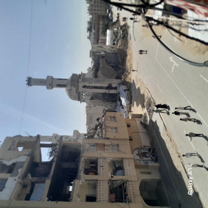 The image displays a severely damaged urban environment in Khan Yunis, Palestinian Territory, under a clear, sunny sky. On the left, a multi-story building exhibits extensive structural collapse on its upper floors, with exposed concrete slabs and rebar. Lower floors show significant damage, including shattered windows and partially collapsed walls, though some balconies still have laundry hanging. In the background, a large mosque with a distinctive minaret and dome stands heavily damaged; large sections of its structure have collapsed, leaving a skeletal appearance and substantial rubble piles around its base, some covered with white tarpaulins.
A paved street runs diagonally across the foreground and middle ground, with unpaved, debris-laden shoulders. A white directional arrow is painted on the road surface. Approximately 15 individuals, primarily men and boys in civilian clothing, are scattered across the street. In the foreground, a group of about seven people are walking or standing, casting long shadows. Further down the street, a pair walks together, and other individuals are dispersed, moving through the scene. The overall scene indicates widespread structural destruction.