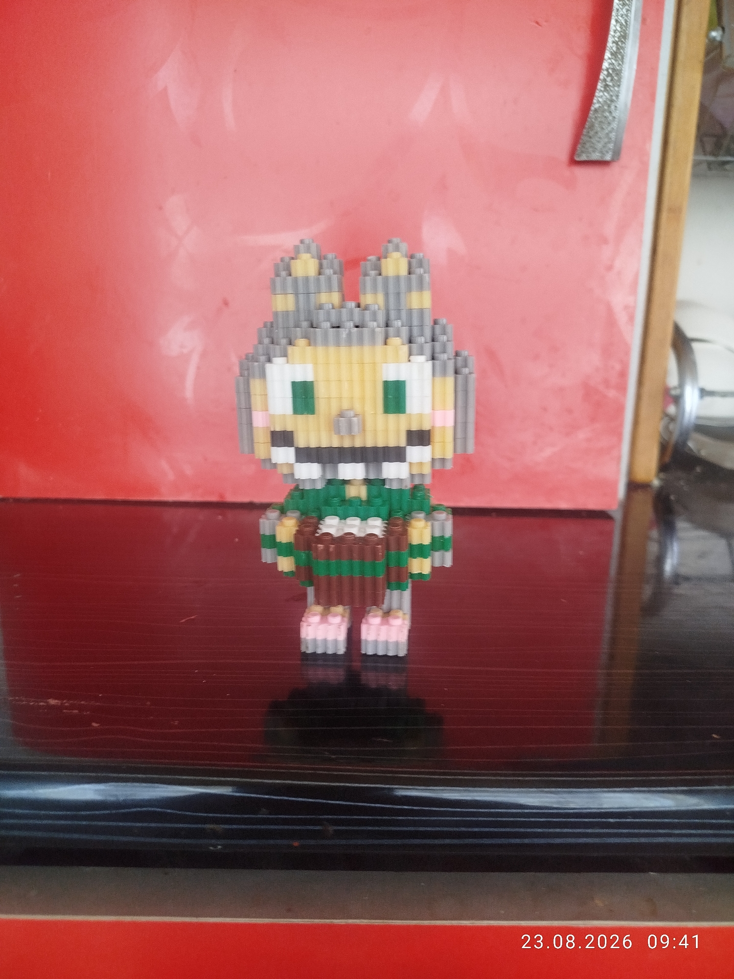 A pixel art figure of a cartoonish character is standing on a dark, reflective surface. The figure is made of small, interlocking blocks and has a light yellow face with green eyes and a wide smile revealing white block teeth. It has gray and yellow block hair and ears, and a green and brown striped body. The figure's feet are pink. The background is a bright red surface, possibly a wall or a backdrop, with some subtle white smudges. To the right of the figure, a wooden frame and a metallic object are visible. The bottom right corner of the image shows a timestamp: "23.08.2026 09:41". The setting does not provide any specific indicators of its location within the city of Don-Aryk, Kyrgyzstan.
