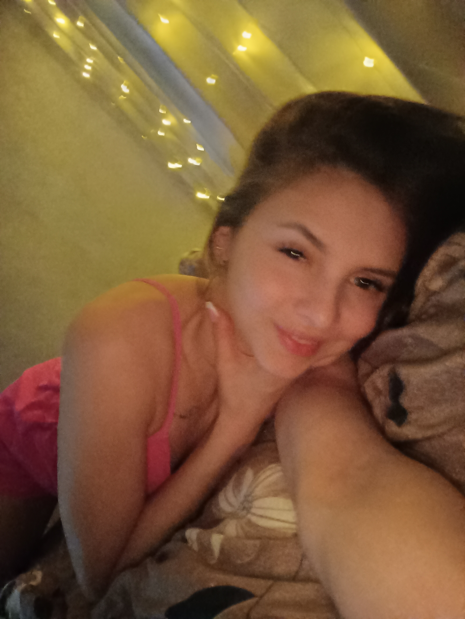 A young woman with brown hair and a pink tank top is lying in bed. She is smiling and her hand is near her face. The background includes a light-colored wall decorated with strings of small, warm-toned lights. The location of this image is not included in this image.