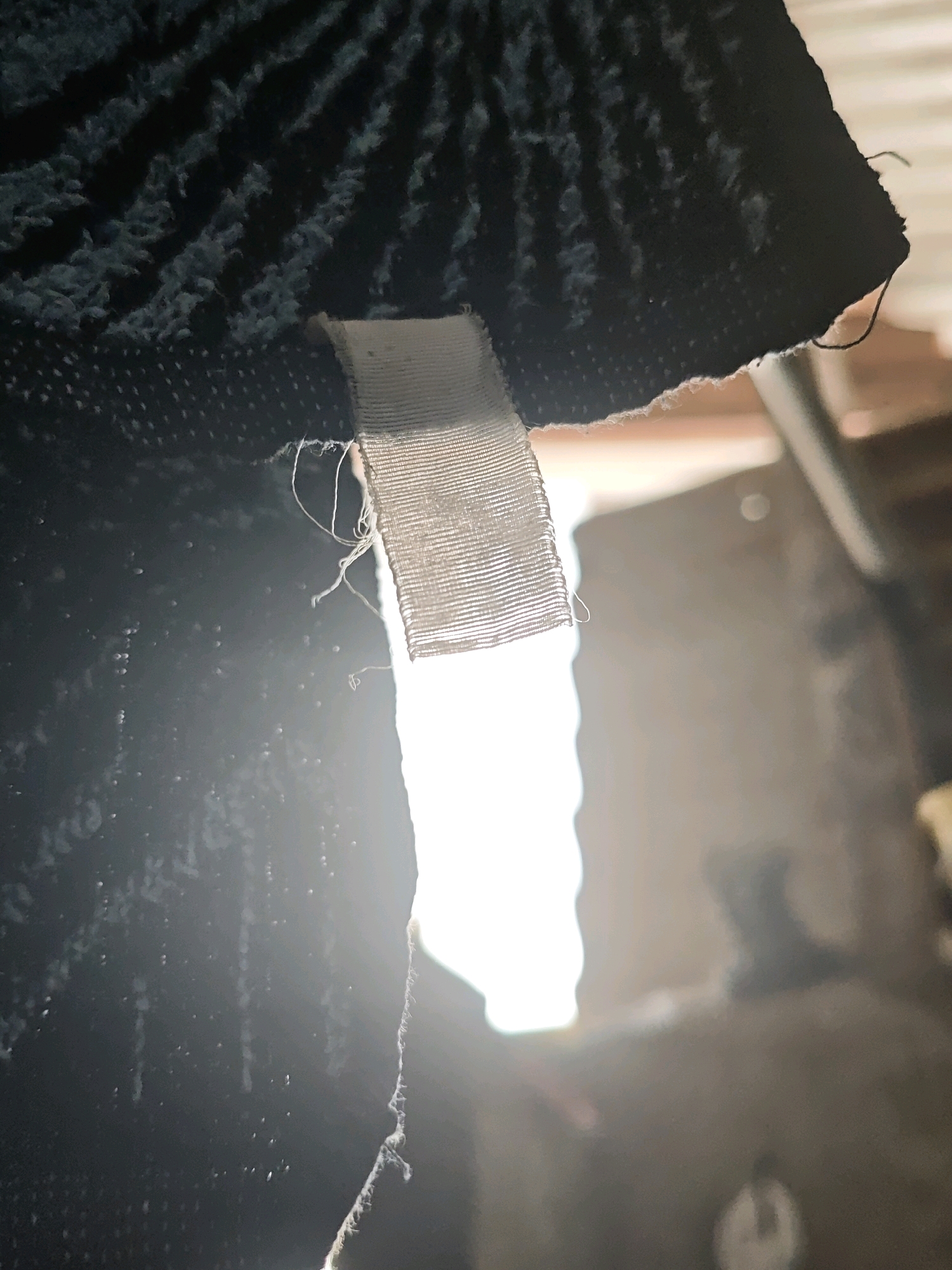 The image is a close-up, low-angle shot of a dark, textured fabric with a white fabric tag attached. The tag is loosely sewn with visible threads. The background is heavily blurred and out of focus, showing light and indistinct shapes that could be part of an indoor environment. There are no identifiable landmarks or features that place this image in Pagu, Indonesia or any specific city.