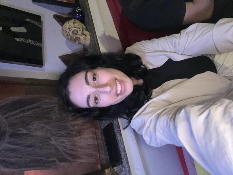 A young adult female with dark, wavy hair and fair skin is depicted from the chest up, smiling directly at the camera. She wears a black top beneath a light-colored, long-sleeved jacket. To her immediate right, a portion of another individual, clad in dark clothing, is visible, suggesting shared seating on a red upholstered surface. The background consists of an indoor environment, likely a cafe or bar. On the left, a dark, textured abstract artwork hangs on a wall, with a black smartphone resting on a light-colored ledge below it. On the right, a light-colored decorative skull with dark etched patterns sits on a similar ledge, alongside a dark blue, white-dotted object and another dark framed artwork mounted above. The specific location within the city of Nha Trang, Vietnam, is not visually identifiable from the image content.