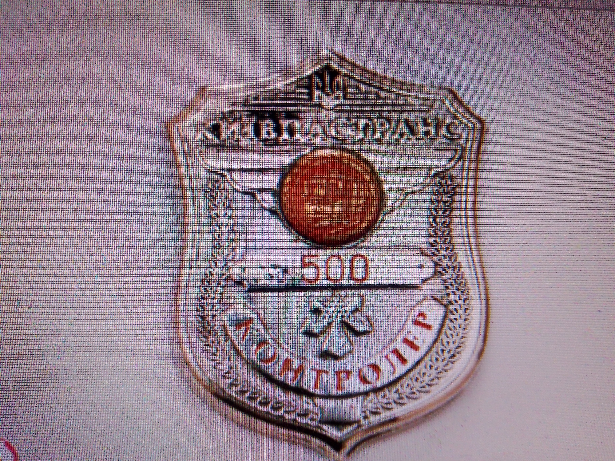 The image displays a close-up, slightly blurry photograph of a metallic badge. The badge is shield-shaped with a stylized emblem at the top and a decorative border. In the center of the badge, there is a red circular insert with an embossed design. Below this, the number "500" is prominently displayed in red text on a silver background. Further down, a red emblem, possibly a stylized flower or star, is visible. The bottom portion of the badge features red text that appears to be in Cyrillic script. The badge has a silver finish with intricate detailing. The background is a light, textured surface, likely a wall or fabric, and appears to be out of focus. No people or specific location details are discernible.