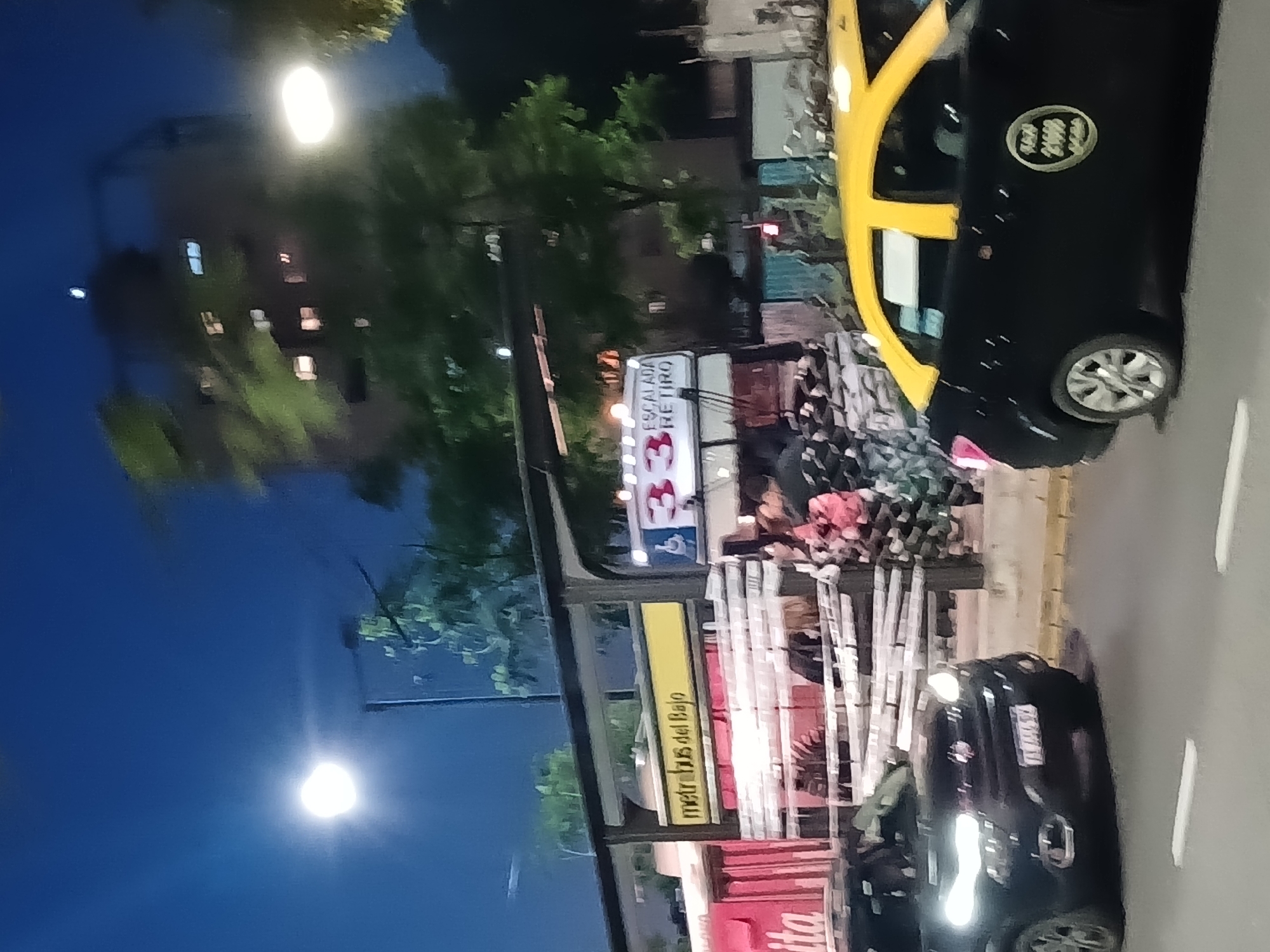 The image depicts a street scene in Buenos Aires, Argentina, at dusk or night. A yellow and black taxi is parked on the right side of the frame. A bus stop with advertisements and signs is visible. The bus stop sign shows "Metrobús del Bajo" in yellow text and "Escalada Retiro" in a white sign on top. A dark-colored car is near the bus stop. A blurred building is illuminated in the background, possibly residential. The sky is dark.