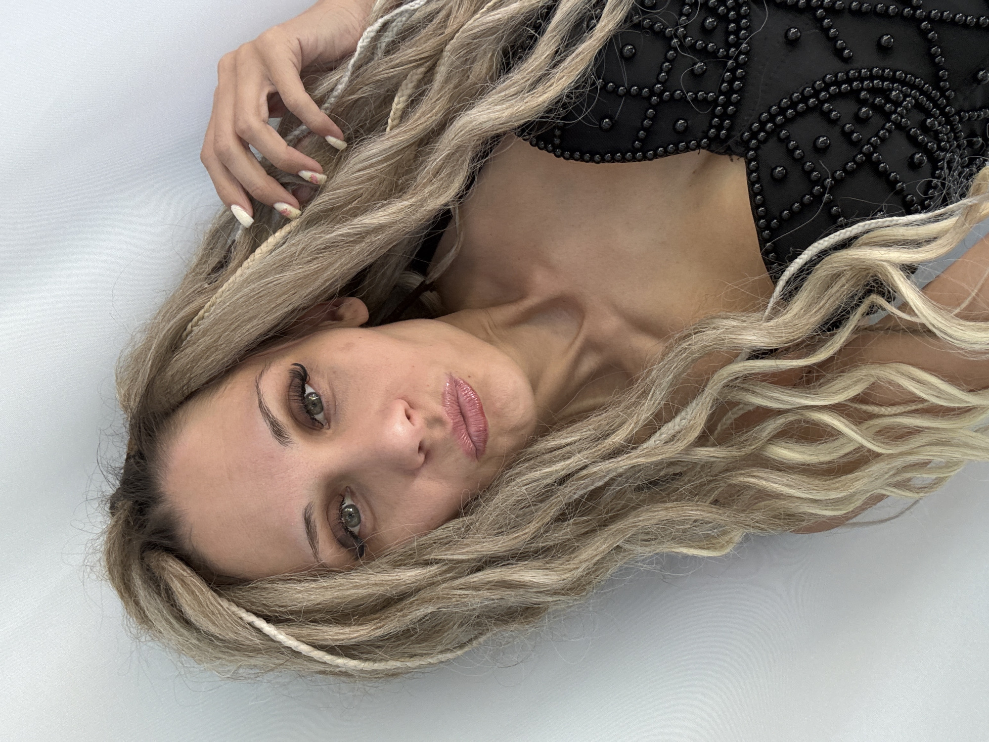 An adult female is depicted in a close-up, head-and-shoulders portrait shot, leaning against a plain, light grey or white textured background, located in the city of Rusanovo, Russia. She has long, blonde hair styled with interwoven lighter-colored braids or dreadlock extensions. Her eyes are light-colored with visible eye makeup, including dark eyeliner and pronounced eyelashes. Her eyebrows are dark and well-defined. Her lips are full, glossy, and slightly pursed in a pout. She is wearing a black garment, possibly a top or dress, featuring a pattern of small, dark, spherical studs or beads across the chest and shoulder area. Her right hand is visible, with light-colored fingernails adorned with subtle pink and white floral or abstract designs, touching her hair near her temple. The lighting is bright and even, illuminating her face and the immediate surroundings.