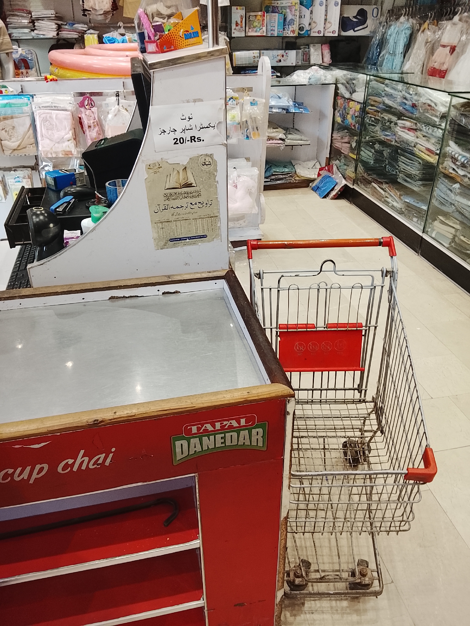 This indoor shot captures a commercial scene, possibly within a store in Gulgasht Colony, Pakistan. The image is dominated by a cash register stand, featuring a surface covered with a metal sheet, and a red base with the "TAPAL DANEDAR" logo. A shopping cart stands adjacent to the cash register, its metal frame showing signs of wear.
Behind the cash register, shelves display various items, possibly clothing, baby products, and other merchandise. Additional shelves and glass display cases with folded items can be seen in the background. A sign is posted near the cash register, written in Urdu, states "Extra shopper charges 20/- Rs"