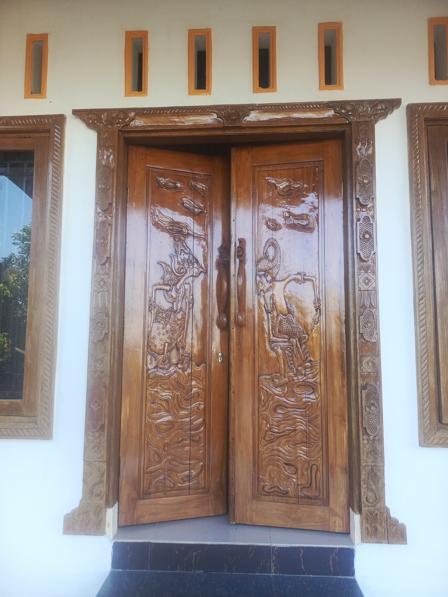 This is a medium-shot, eye-level, outdoor photograph of a wooden double door with intricate carvings. The doors are made of a medium-toned wood with a glossy finish and are slightly ajar. Each door panel features a detailed carving of a traditional Indonesian puppet figure, possibly from a Wayang performance, set against a background of swirling water or waves. The figures appear to be male, adorned with elaborate headdresses and traditional clothing. The door frame is also made of wood and features ornate carvings, including floral motifs and geometric patterns. Above the doors, on the white wall of the building, are three narrow, rectangular vents, each with an orange frame. To the left and right of the doors are single windows with wooden frames. The window on the left reveals greenery and trees outside. The ground in front of the door appears to be a tiled step with dark, possibly marble or granite, tiles. The overall style suggests a traditional Indonesian architectural aesthetic. The location is identified as Kendari, Indonesia, implying the photograph was taken at a residence or public building in that city. There are no people or animals visible in the image.