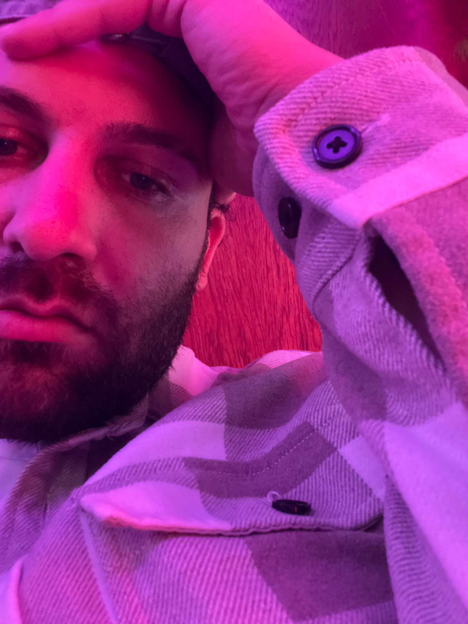 A close-up selfie shows a person's face and torso, illuminated by pink and purple lighting. The individual has dark hair and a beard, with one hand resting on their forehead. They are wearing a plaid shirt with a button visible on the cuff. The background appears to be a wooden surface. The image does not provide any information to determine its location in the city of Newry, United Kingdom.