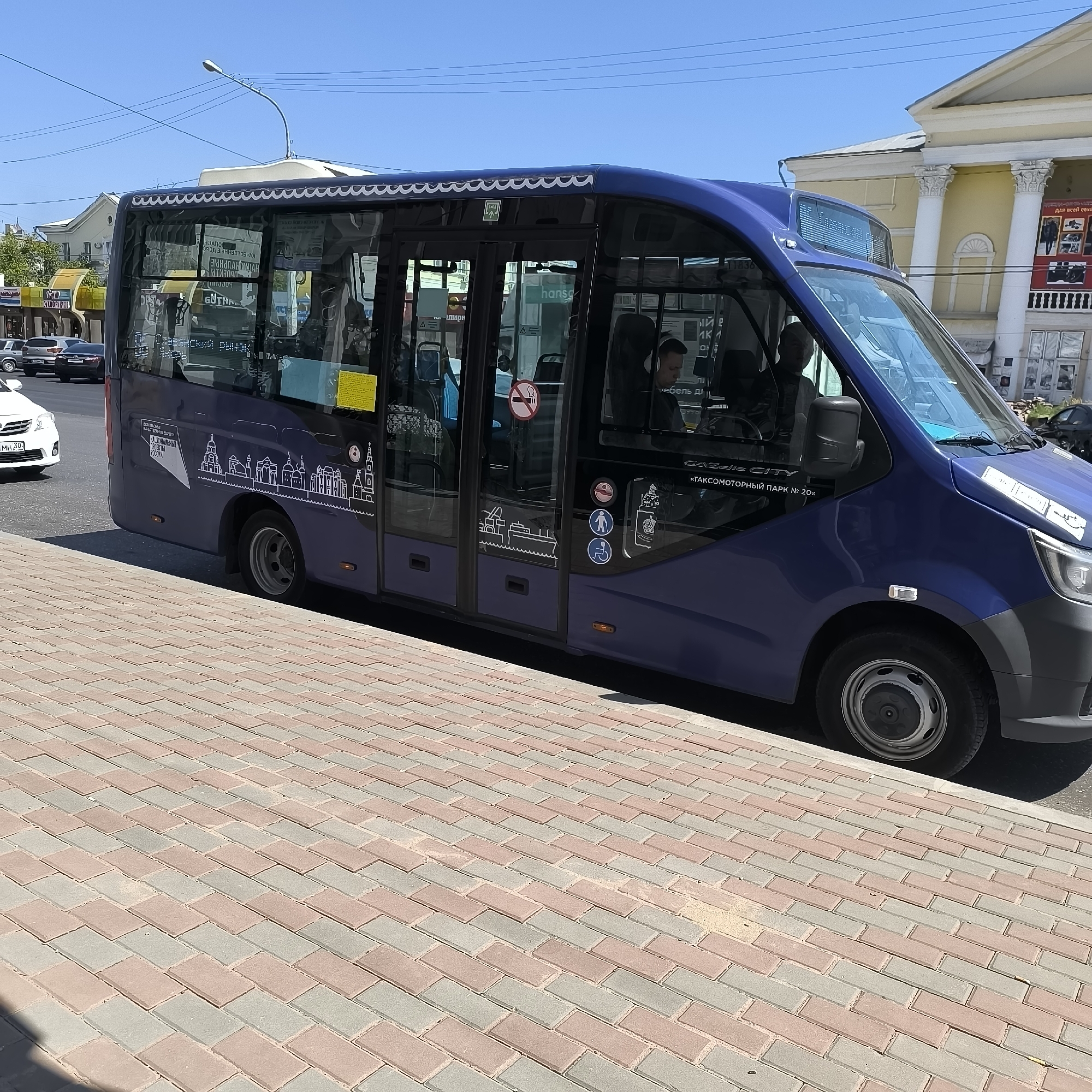 A dark blue minibus, identified by the text "GAZELLE CITY" and "ТАКСОМОТОРНЫЙ ПАРК № 20»" on its side, is parked on a street in Astrakhan, Russia. The minibus features a decorative white wavy trim along its roofline and line drawings of city buildings and a ship on its side. The bus has tinted windows and a driver is visible inside. In the background, a building with classical columns and yellow facade stands, possibly a cultural or administrative building. A street lamp with a curved arm is positioned above the bus. The foreground shows a pedestrian walkway paved with interlocking bricks in alternating shades of red and gray. Several other vehicles, including a white car and a dark sedan, are visible on the road behind the bus. The sky is clear and blue, indicating a sunny day.