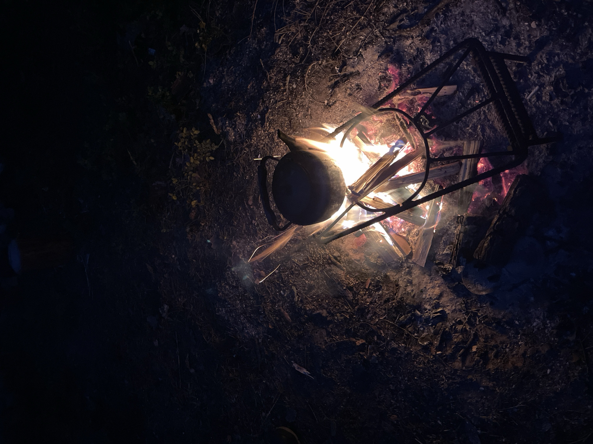This is a photograph taken at night, likely in an outdoor setting. A bonfire is burning in the foreground, with visible flames and glowing embers. A metal kettle is suspended over the fire, supported by a wire rack. The ground surrounding the fire appears to be covered in dirt, leaves, and twigs. The overall lighting is dim, with the fire providing the primary light source. No people are visible in the image, and no specific landmarks or urban elements can be identified to pinpoint a location in Roshchino, Russia.