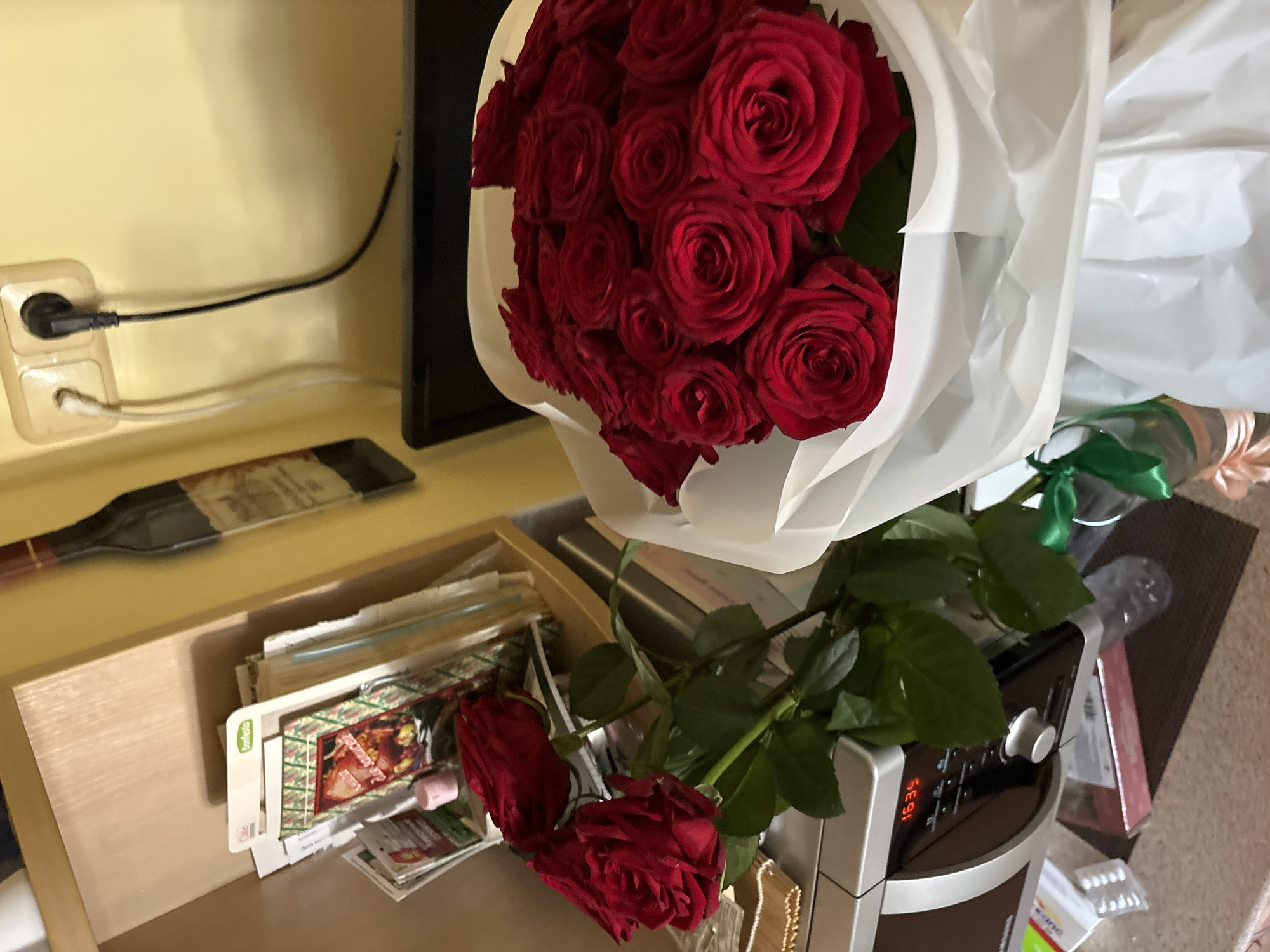 The image depicts a still life of a large bouquet of red roses presented in white paper. Some roses are loose on a metallic surface in the foreground, while others are clustered in the bouquet. Behind the roses, a microwave oven displays the time "16:39". A shelf unit is visible to the left, holding various items including what appears to be boxes of food and possibly recipe cards. The background features a yellow wall with an electrical outlet and plug. To the left of the outlet, a decorative wine bottle is mounted on the wall. The overall setting suggests an indoor environment, possibly a kitchen or living space. The image is taken from a slightly elevated angle, looking down at the arrangement. There are no people present in the scene. The lighting is warm, suggesting interior lighting. The specific location is identified as Minsk, Belarus.