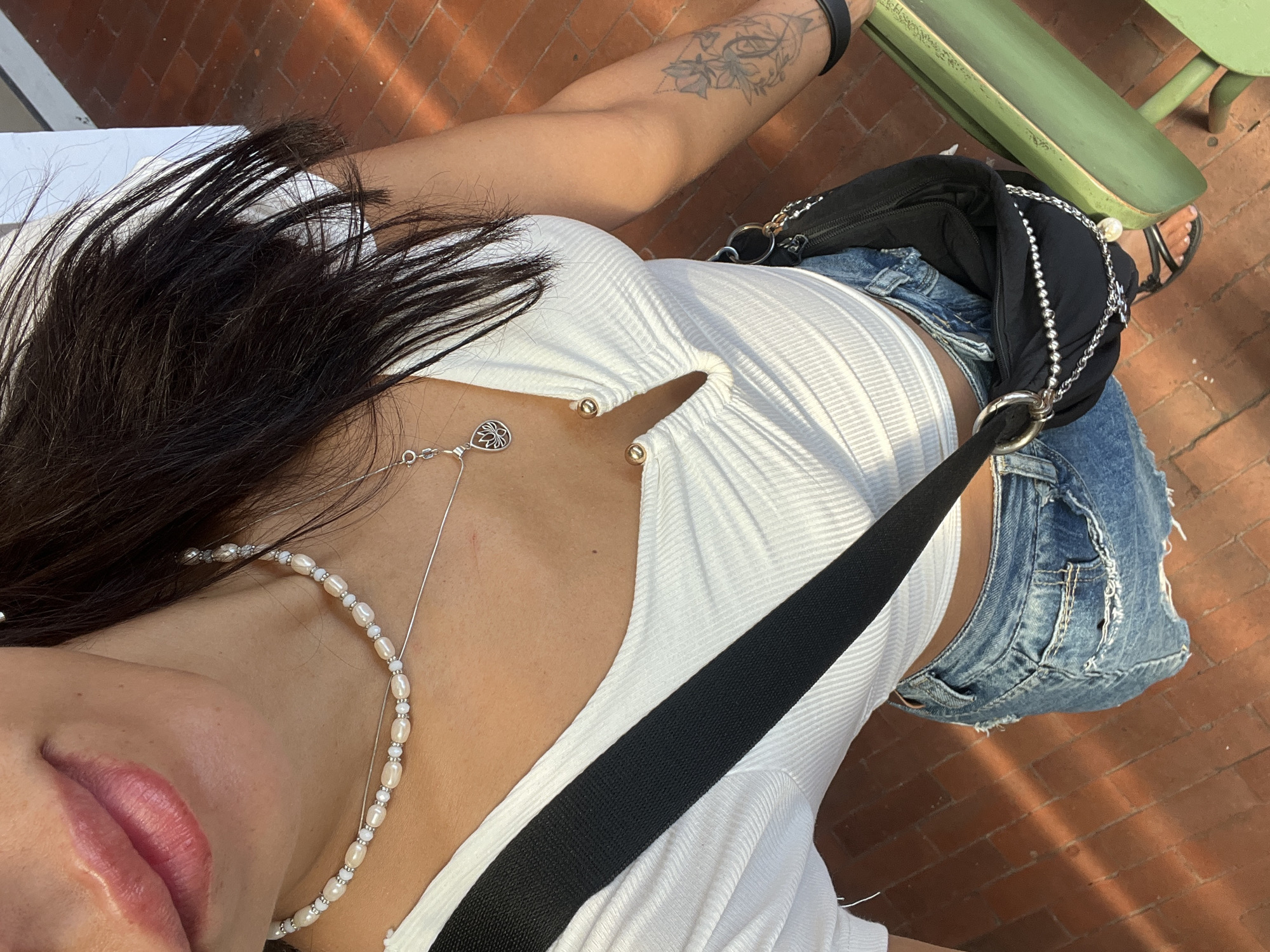 A downward-angled shot captures a partial view of a person seated on a green bench on a brick-paved surface. The individual is wearing a white ribbed top, faded and distressed denim shorts, and a black handbag with a silver chain detail. A black strap cuts diagonally across their torso. The visible arm has a floral tattoo. The location is assumed to be Kuta Selatan, Indonesia, based on the prompt. The scene lacks explicit context to confirm any actions or interactions.