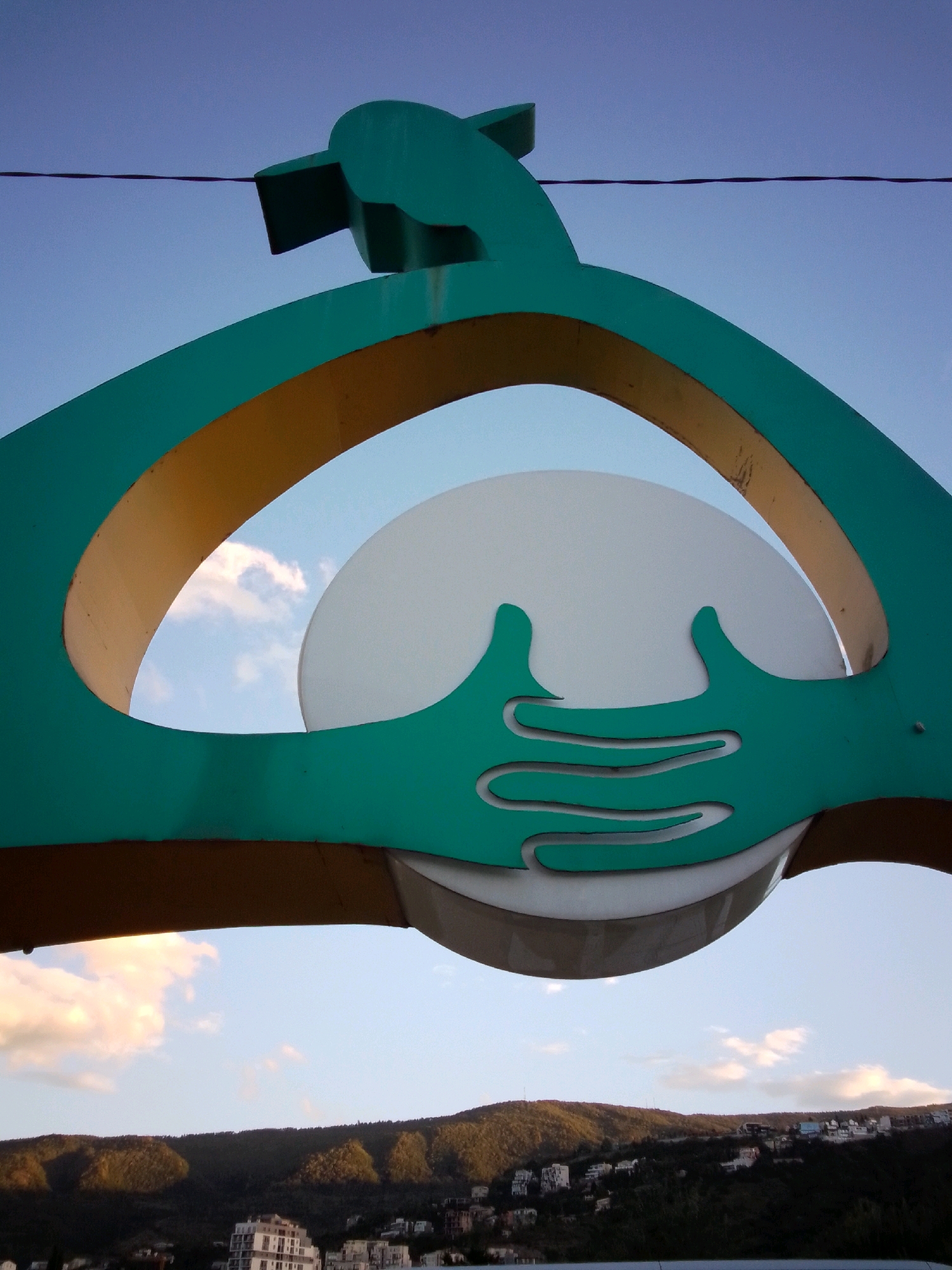 A low-angle outdoor photograph shows a large, teal-colored abstract sculpture against a clear blue sky. The sculpture appears to be made of metal and is shaped like two stylized hands cradling a white disc. A thin, dark wire runs horizontally across the top of the frame, crossing in front of the sculpture. Below the sculpture, a distant, hilly landscape is visible, dotted with buildings and trees. The light suggests it is either early morning or late afternoon. The sculpture's location is unclear and cannot be determined from the image alone.