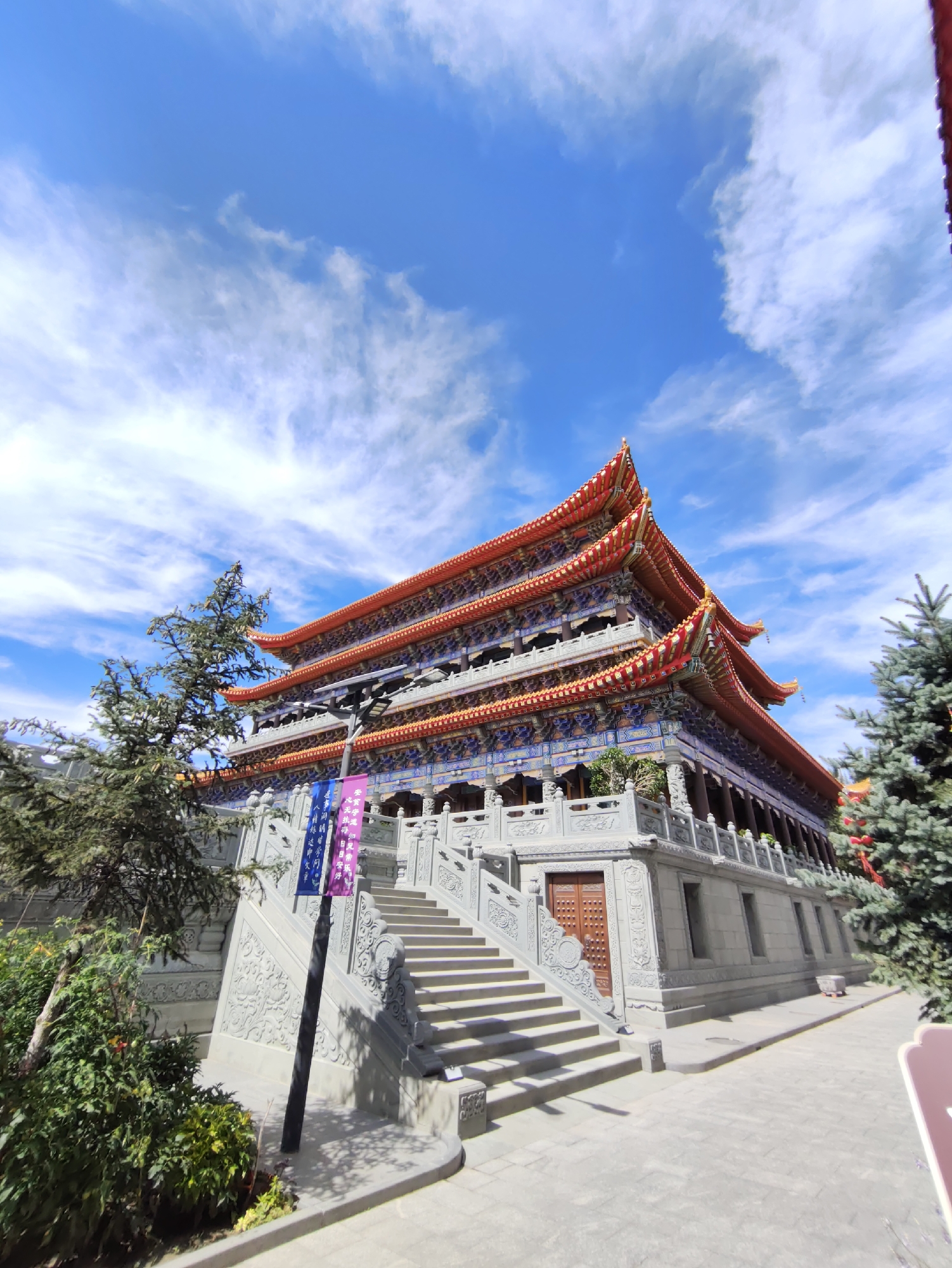 This is a wide-angle, eye-level shot of a multi-tiered Buddhist temple under a bright blue sky with wispy white clouds. The temple features an ornate, brightly colored roof with red and gold accents and intricate blue and red decorative elements along its eaves and facades. A wide, stone staircase with decorative carvings leads up to the main entrance, which has a wooden door. The temple is surrounded by lush green trees and shrubs. Banners hang from a lamppost near the stairs. The ground in front of the temple is paved with light-colored stones. The overall impression is one of grandeur and spiritual significance, located in the city of Daqing, China.
