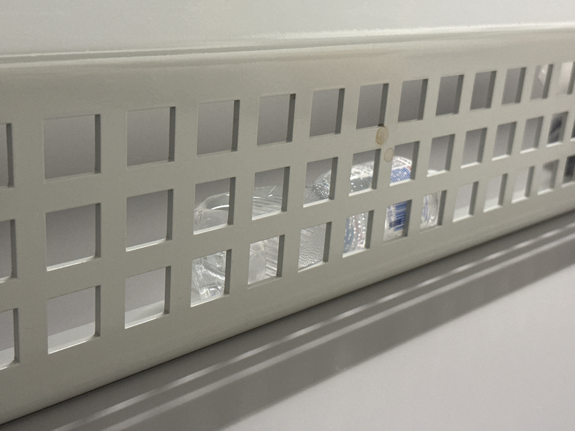 The image shows a close-up, low-angle shot of a white, rectangular grille with a grid of square openings. Behind the grille, several clear plastic bottles are visible, some containing liquid. The bottles are unevenly distributed behind the openings. A faint shadow is cast on the left side of the image by an unseen object. The overall scene suggests an interior environment, possibly a storage area or a commercial setting.