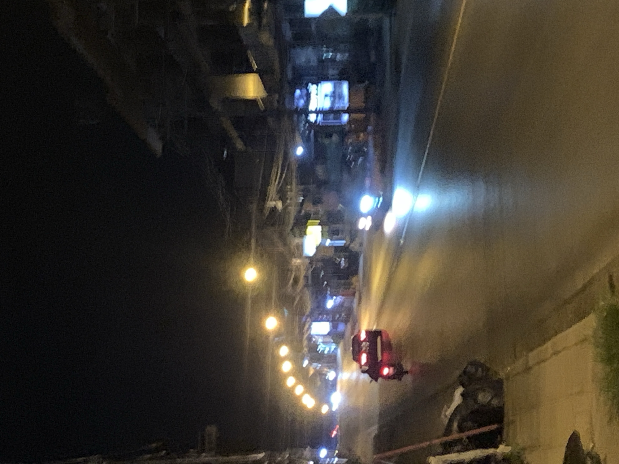 The image depicts a nighttime scene in บ้านบางรักษ์, Thailand. A road runs vertically through the right side of the image. On the right side of the road, there is a low wall with some greenery. On the left side of the road, there's a row of buildings, some illuminated by lights. Streetlights line the side of the road. A vehicle with red lights is seen on the road. In the bottom right corner, there is a person or a group of people, possibly seated. The left side of the image is mostly dark.