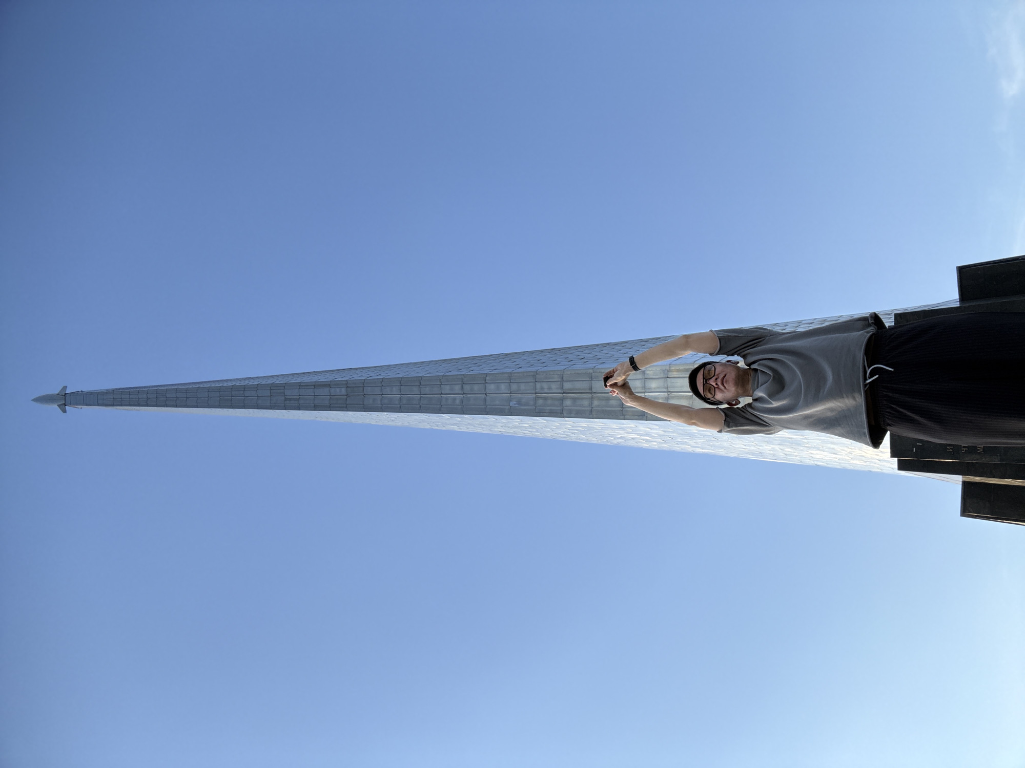 A low-angle, full-shot image shows a person standing at the base of the Monument to the Conquerors of Space, a tall, obelisk-shaped monument topped with a rocket. The monument is located in Moscow, Russia, and is made of titanium and stainless steel, with a mosaic of inlaid panels. The sky is clear and blue. The person, a young male wearing a grey t-shirt and black sweatpants, stands with arms raised above his head, as if to embrace the monument. He wears glasses and a watch on his left wrist. The monument's base is made of dark stone, and there are inscriptions on the stone panels.