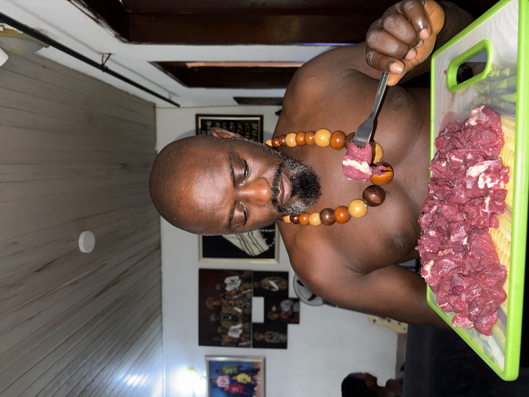A dark-skinned, bald man with a grey beard and wearing a wooden beaded necklace is shown in a close-up, slightly tilted portrait. He is holding a fork with a piece of raw meat on it. Next to him is a green-bordered cutting board piled with more raw meat. The background shows a room with wood-paneled walls and framed pictures on the wall. The image appears to be taken indoors, possibly a residence in Ado, Nigeria.