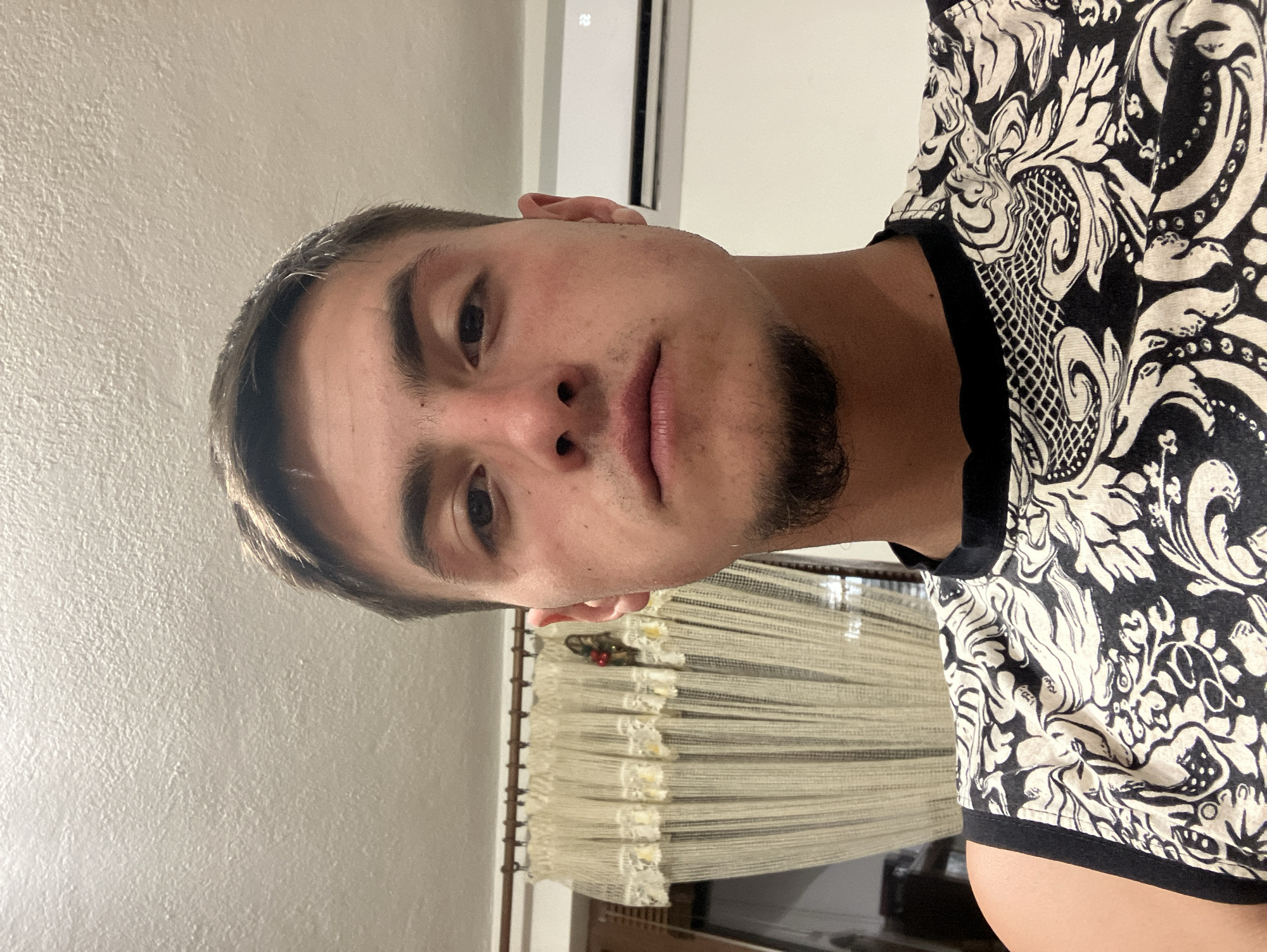 A close-up, indoor photograph features a young man with light skin and short brown hair, facing the camera with a neutral expression. He has a stubble beard and light-colored eyes. He is wearing a patterned black and white shirt with a black trim at the neck.
The background includes a light-colored wall and a window with cream-colored horizontal blinds. A portion of what appears to be an air conditioning unit is visible at the top of the frame.
There is no information about this image being located in Krastevich, Bulgaria.