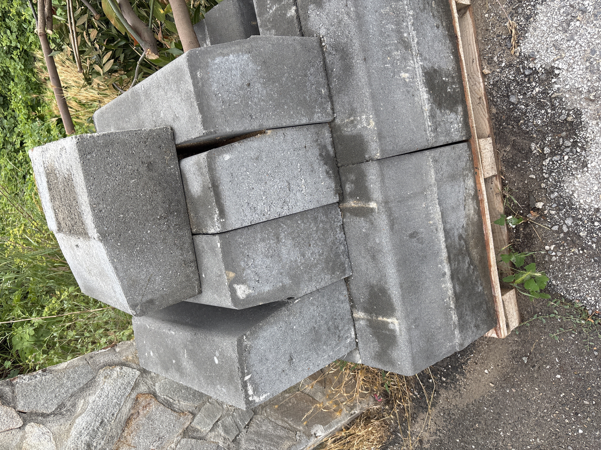 A pile of concrete curbing blocks is stacked on a wooden pallet. The blocks are grey and appear to be used, with some dirt and water stains on their surfaces. They are arranged in a haphazard manner, with some leaning against each other. To the left of the blocks is a stone wall, constructed from irregular grey and brown stones. Behind the blocks and wall, there is dense green foliage and some dry grass. A few branches are visible in the upper right portion of the image, extending from a tree. The ground in front of the pallet is a mix of asphalt and dirt, with small pebbles and some green weeds growing through. The scene appears to be outdoors, possibly in a construction or landscaping area within the village of Garmen, Bulgaria.