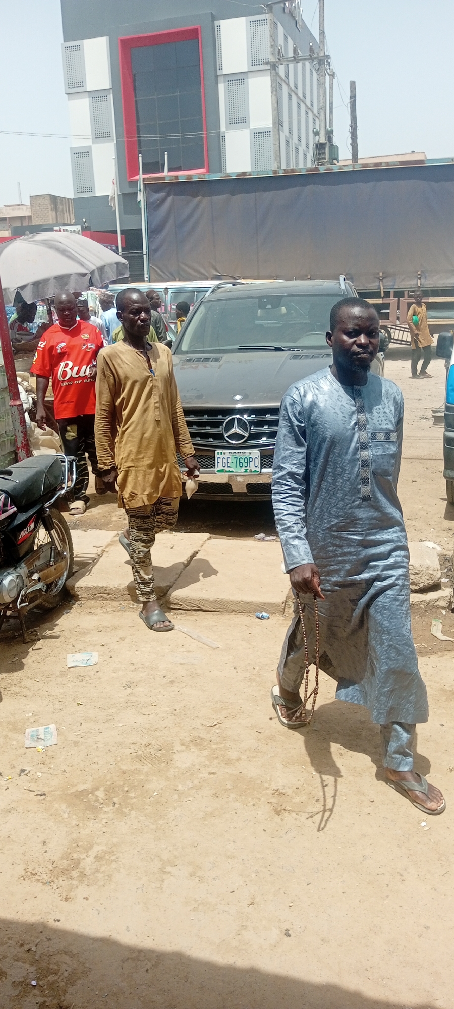 A medium shot captures a street scene in Fagge C, Nigeria. In the foreground, two men walk on a dirt and gravel path. The man on the right wears a blue patterned robe and holds a string of prayer beads in his right hand. He has a beard and short black hair. The man on the left wears a tan, long-sleeved tunic and patterned trousers. He has a shaved head and appears to be walking. Behind them, a dark-colored Mercedes-Benz SUV is parked, its license plate visible as "FGE-769PC KANO". To the left of the SUV, a man in a red t-shirt with "Budweiser" printed on it walks past. A motorcycle is parked to the far left of the frame. In the background, a modern building with a grey facade, white accents, and a large red-trimmed window is visible, along with a large truck with a dark tarp. The environment appears to be an urban outdoor setting with a sunny sky.