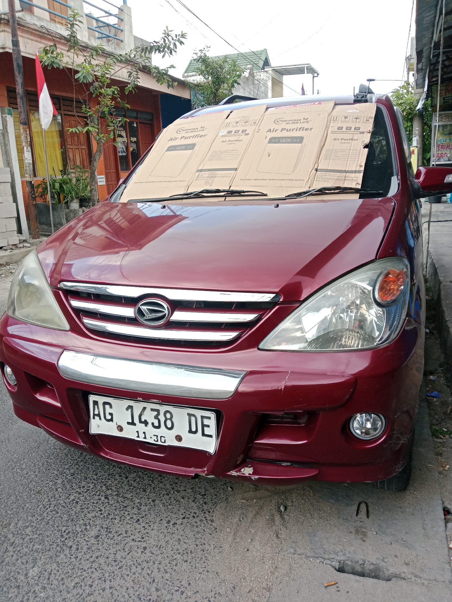 The image shows a dark red Daihatsu car parked on the side of a street in Kediri City, Indonesia. The car's windshield is covered with large cardboard pieces advertising "Air Purifier" from "ONE HOME MISS LIFE". A red and white Indonesian flag is visible on a pole to the left of the car, next to a building with wooden doors. The car has a license plate reading "AG 1438 DE". The street is paved with asphalt, and a concrete sidewalk runs alongside it. The environment appears to be a commercial or residential area, with buildings and trees visible in the background. There are no people in the image.