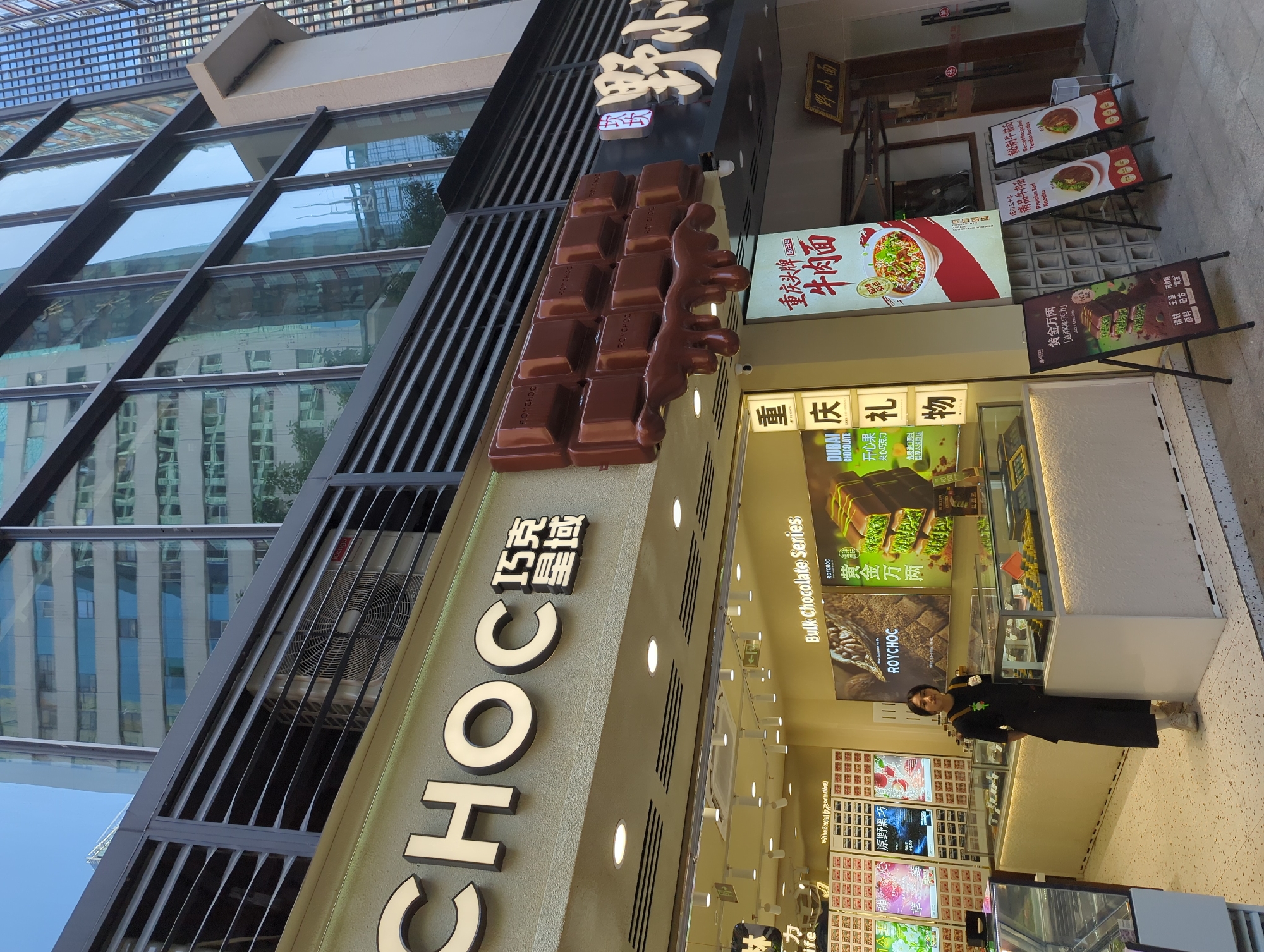 The exterior of a chocolate shop named "CHOC" is captured on a sunny day in Yuzhong District, China. The shop's facade features a large, illuminated sign with the English word "CHOC" and Chinese characters above. A prominent 3D chocolate bar sculpture is mounted on the building. Below the signs, the shop's interior is visible, displaying refrigerated display cases filled with chocolates and other sweets. A young person wearing a black apron and beige pants stands in front of one of the display cases, facing the camera. Several A-frame signs are placed on the sidewalk outside the shop, advertising "Premium Beef Noodles" and a chocolate product. The background shows modern, tall buildings with reflective glass windows, indicating an urban environment. The overall scene suggests a commercial street in a city.