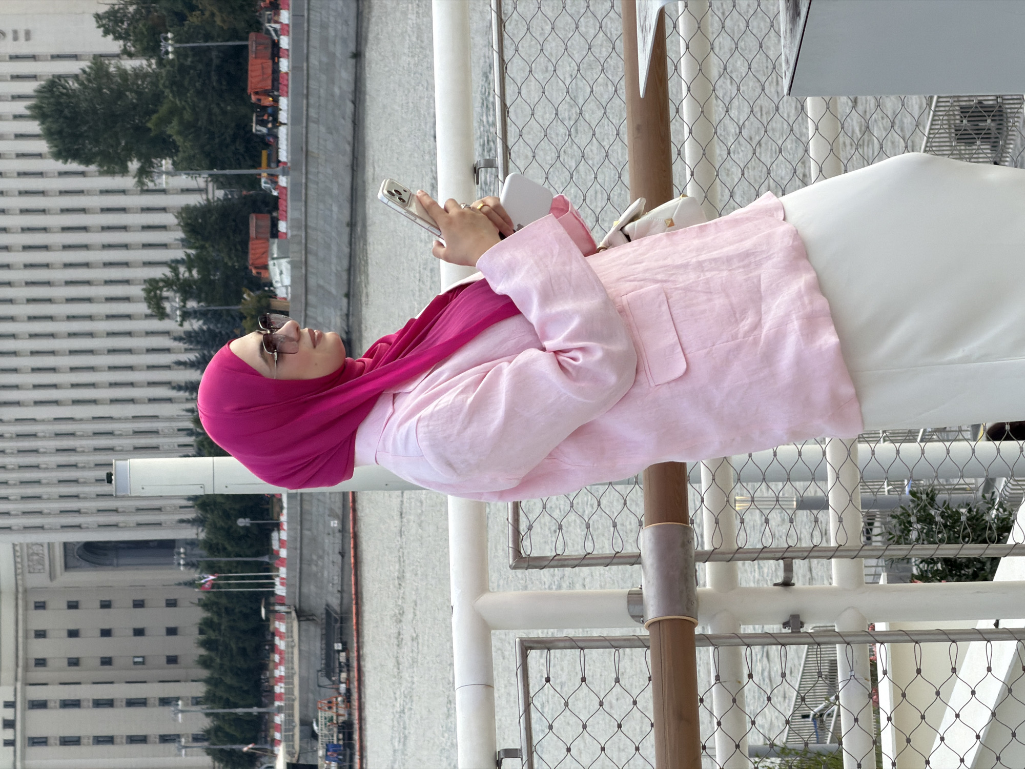 A woman wearing a pink hijab and a pink linen blazer stands on a pedestrian walkway, looking at her phone. She is positioned in profile, facing right. The woman is wearing a white skirt and sunglasses. In the background, a large, ornate building with many windows and an arched entrance is visible. Behind the building, trees and trucks can be seen. The walkway has a metal railing with a wooden top. A body of water, likely a river, is visible beyond the railing. The architecture of the building suggests it is a significant structure in Moscow, Russia, possibly an administrative building or a university.