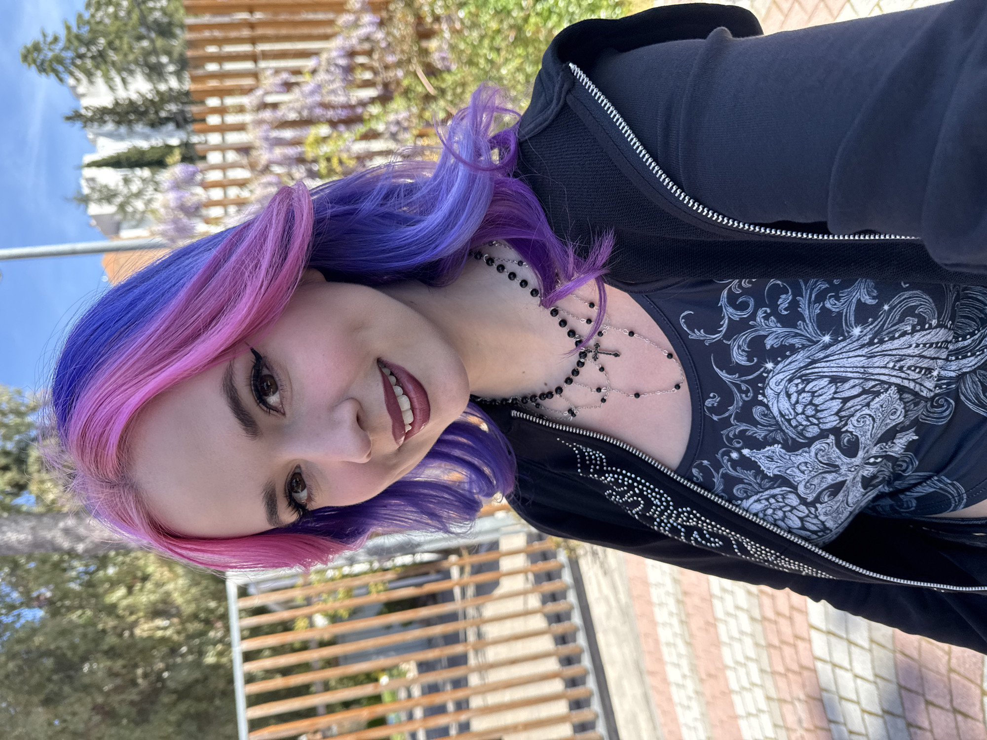 A woman with dyed purple and pink hair, wearing a black hoodie over a patterned black top and a black beaded necklace, is taking a selfie. The background includes a wooden structure, possibly a pergola, with blooming purple wisteria. There are trees visible in the distance. The location appears to be outdoors, possibly a park. The presence of wisteria and certain architectural features suggests a Mediterranean environment, potentially consistent with Madrid, Spain, but the image does not offer definitive indicators that would point towards a specific neighborhood in the city.
