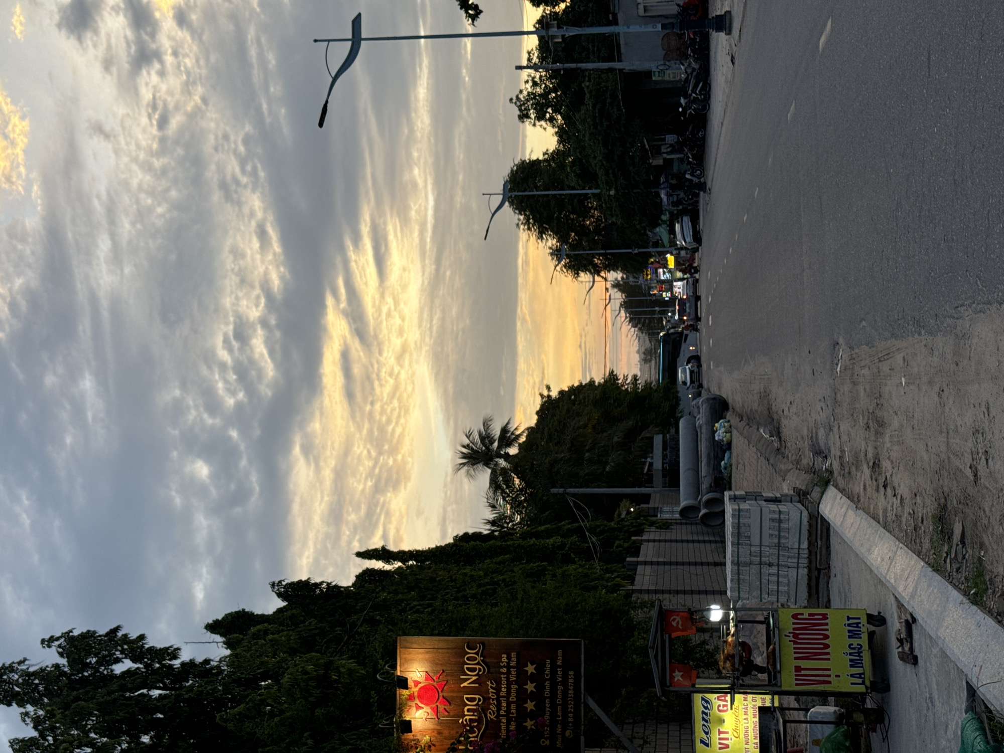 The image shows a street scene in Mui Ne, Vietnam at dusk. The sky is filled with dramatic clouds illuminated by the setting sun, casting a warm glow. On the left side of the road, a tall sign for "Hoàng Ngọc Resort" is visible, with a stylized sun logo and text in Vietnamese. Below the resort sign, a food cart selling "Vit Nuong" (grilled duck) is set up, with a whole roasted duck on display. Next to the food cart are stacks of grey paving stones and large pipes. The road itself is paved and appears to be a main thoroughfare, with several cars, motorbikes, and a bus visible in the distance. Streetlights line the road, some of which are illuminated. Trees and lush foliage line the left side of the street, partially obscuring buildings behind them. The overall impression is a bustling yet tranquil evening scene in a coastal Vietnamese town.