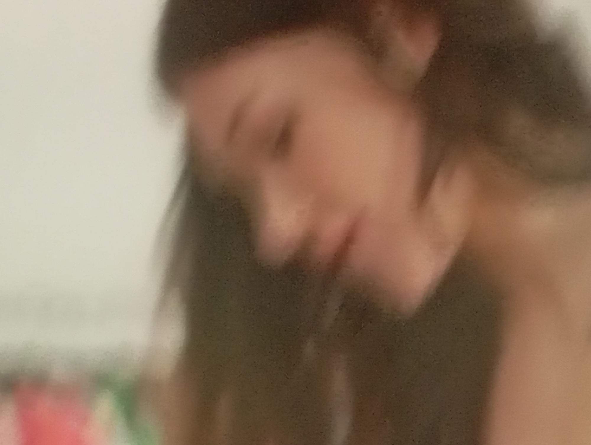 The image is a close-up, blurry photograph of a person's face, predominantly the left side of their face and hair. The person appears to be looking downwards and to the left. The background is a plain, light color. The lighting is soft and diffused. There are no discernible objects or environmental details indicative of a specific city or location.