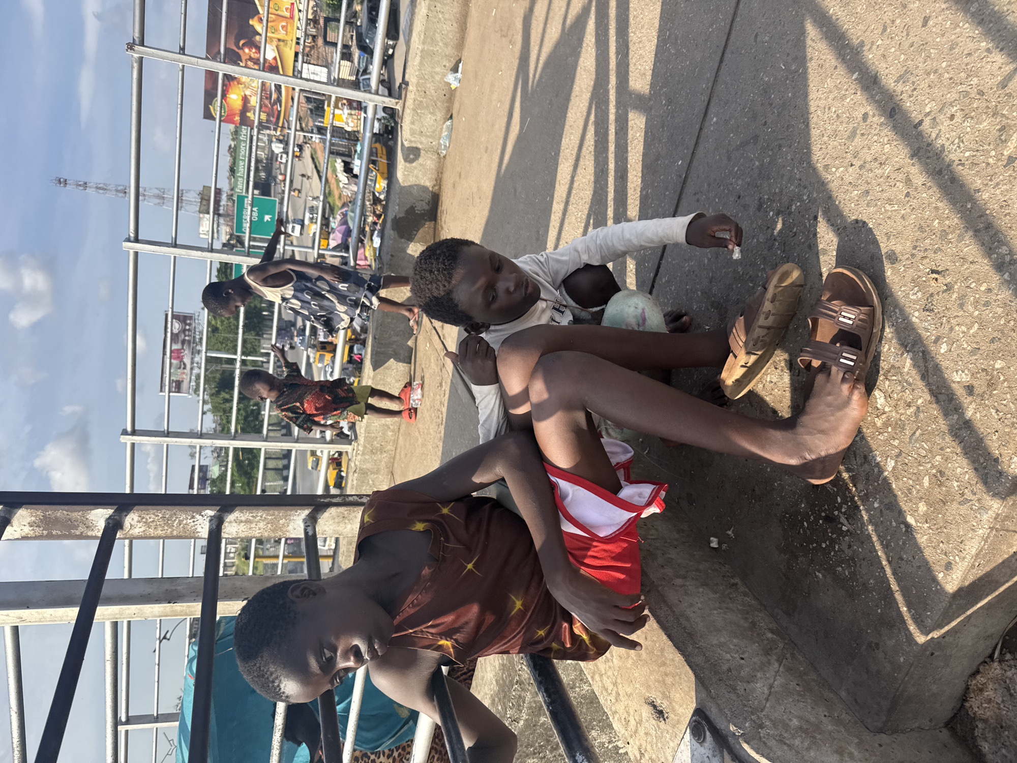 The image captures a scene in Onitsha, Nigeria, likely on a bridge or overpass overlooking a busy street. In the foreground, two young boys are the primary subjects. The boy on the left, sitting with his legs extended, wears a brown sleeveless shirt with a star pattern and red and white shorts. He is looking off to his left, with a somewhat pensive expression. To his right and slightly behind him, another boy is crouched down, wearing a white long-sleeved shirt and light-colored shorts. He is holding a small white object, possibly a piece of candy or a cigarette, between his fingers, and appears to be examining it. His feet are bare, and he is wearing brown sandals.
In the background, several other children are visible, with one child in a colorful patterned shirt standing and another in a dark and white patterned dress walking away from the camera. The environment suggests an urban setting with a road below, filled with various vehicles including yellow minibuses, cars, and trucks. Structures and billboards are also present, indicating commercial activity. The sky is partly cloudy, and the sun casts distinct shadows across the concrete ground. The overall scene depicts a moment of respite or interaction among children in a bustling city environment.