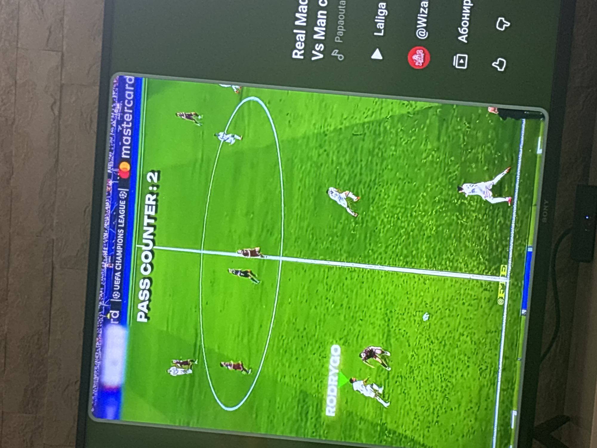 The image displays a television screen in an indoor setting, viewed at a slight upward angle. The screen shows a split display: a football game on the left and a YouTube interface on the right.
On the left side, a simulated football match is in progress on a green pitch with white markings, including a center circle and a halfway line. The top banner of the football display includes "UEFA CHAMPIONS LEAGUE" text and a "mastercard" logo. Below this, an overlay reads "PASS COUNTER: 2". Multiple animated players are visible, some wearing white jerseys and others burgundy or dark-colored jerseys. One player in a white jersey, located in the lower-left quadrant, has the name "RODRYGO" displayed above him. A football is situated near the center of the pitch.
The right side of the screen presents a YouTube interface. A video title, partially visible, reads "Real Mad Vs Man c" (likely "Real Madrid vs Man City"). Below the title, a music note icon precedes "Papaouta", and a play icon precedes "Laliga". A circular red channel icon with a white logo is next to "@Wizar". Further down, a button in Cyrillic reads "Абонир" (Subscribe), followed by thumbs-up and thumbs-down icons for liking or disliking the content.
The television set itself is a SONY brand, indicated by the logo at the bottom center of the screen's frame. Behind the television is a textured wall with a pattern resembling light-colored bricks or stone. A black cable runs horizontally below the television, and a dark rectangular electronic device is partially visible on the surface supporting the TV.