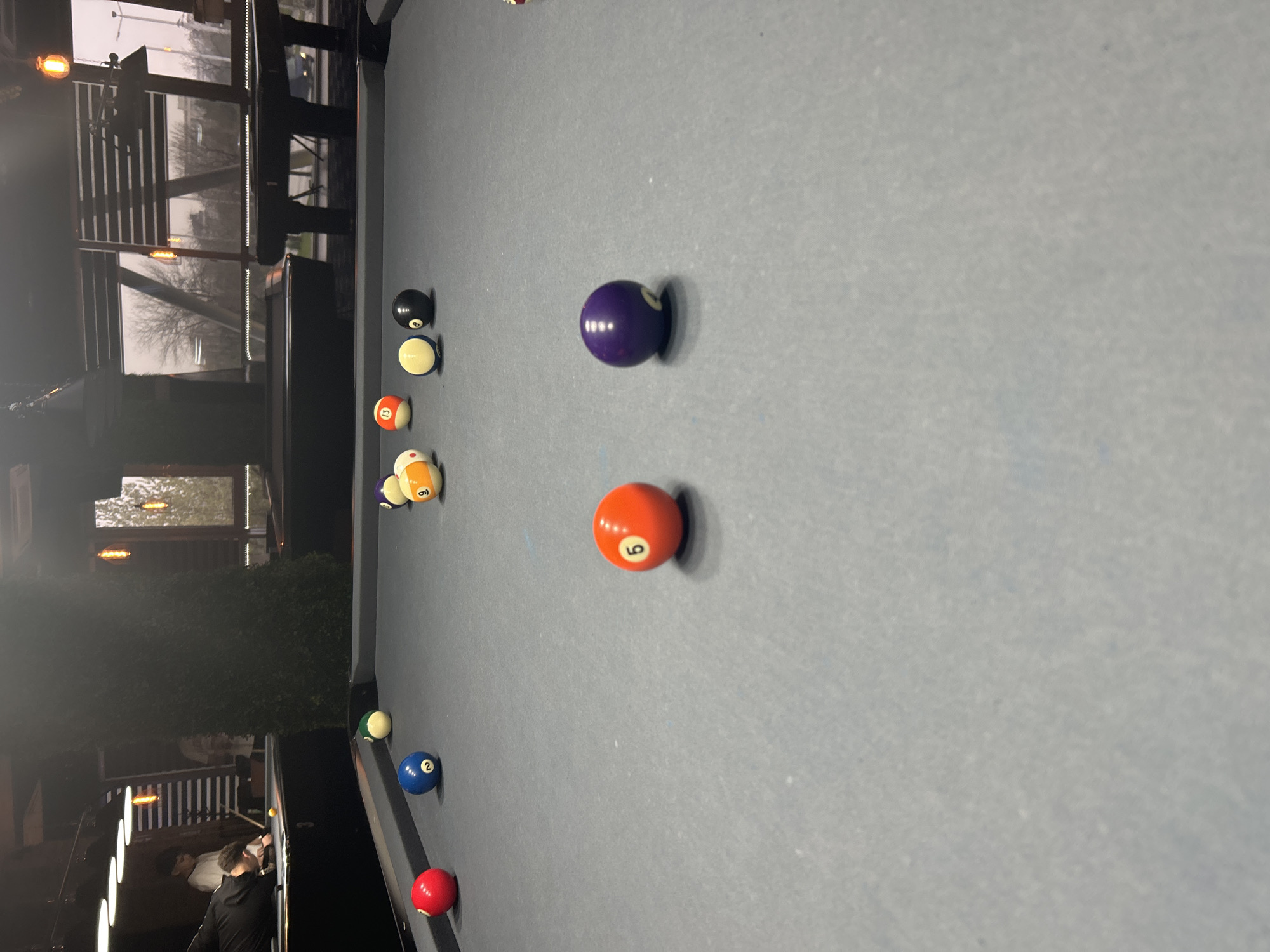 The image depicts a pool table in an indoor setting, possibly a bar or entertainment area, located in Plovdiv, Bulgaria. Several pool balls are scattered across the table: a purple 4-ball, an orange 9-ball, a black 8-ball, a yellow ball, a red 5-ball, a striped 15-ball, a green 2-ball, and a red ball. A person is visible at the edge of the table, holding a pool cue, suggesting they are about to take a shot. Outside the window, foliage and building details can be seen. The table's surface is gray.