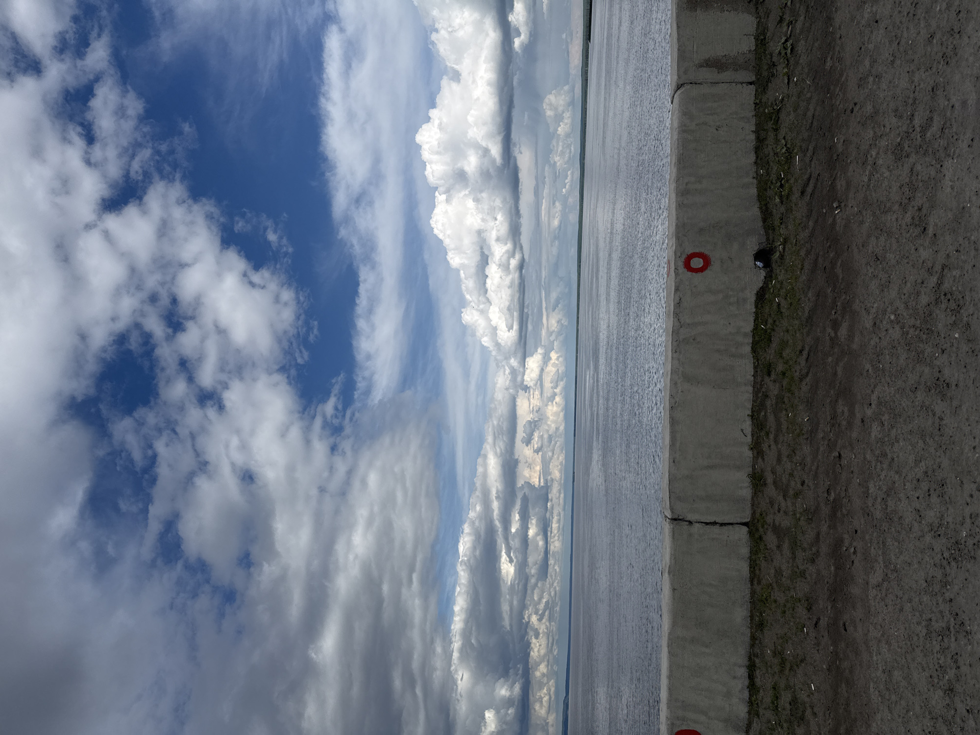 A sideways view of the Kama Reservoir, taken from the city of Chaikovsky, Russia. The image features a body of water, likely the reservoir, stretching horizontally across the middle. To the left, the sky is filled with billowing cumulus clouds, reflecting in the water. To the right of the water is a grey curb with red markers and a black bird on top, adjacent to a dirt path.