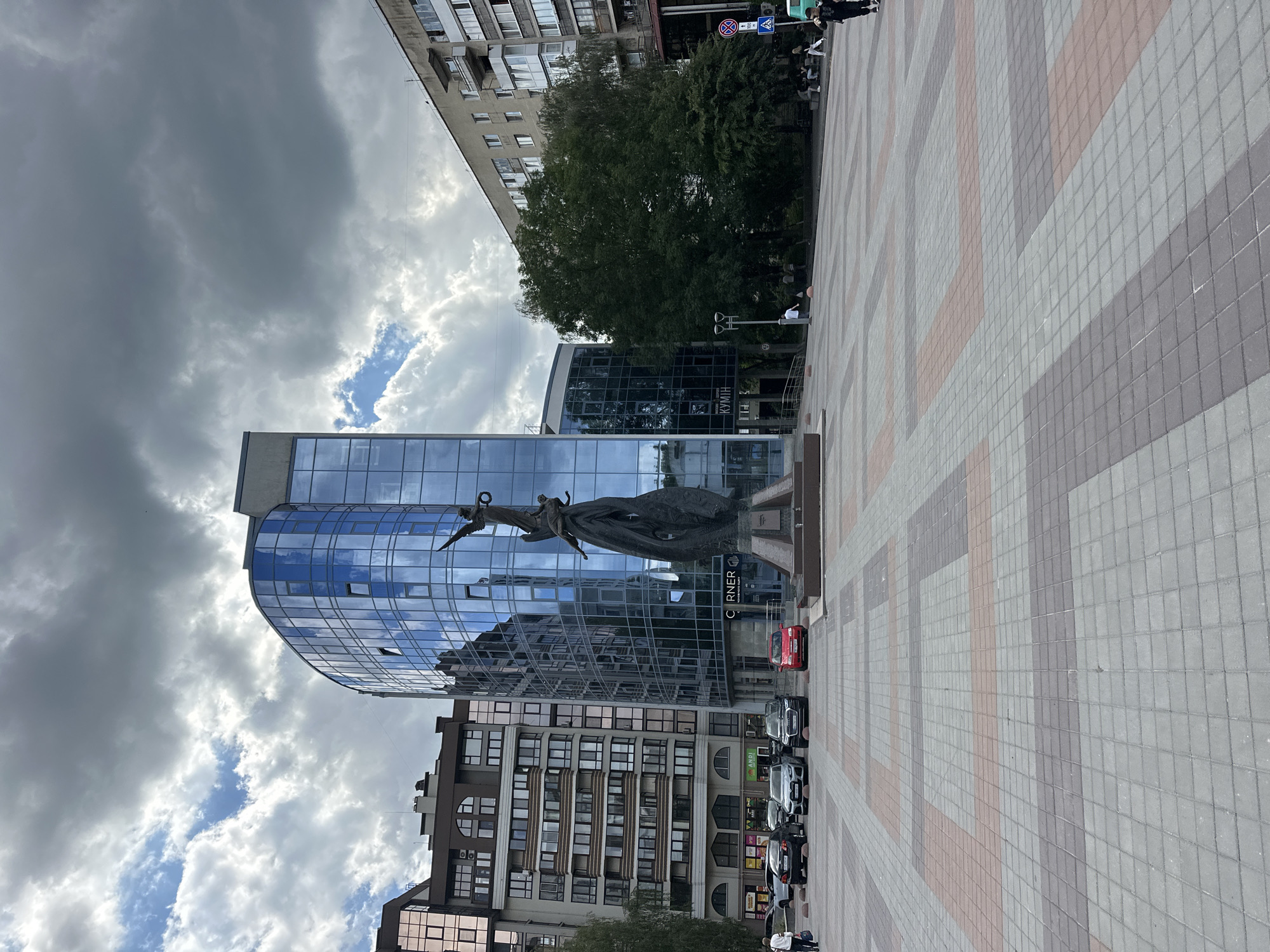 In Ternopil, Ukraine, an outdoor plaza is depicted on a cloudy day. In the center of the frame stands a dark, abstract sculpture featuring two figures reaching upwards. Behind the sculpture is a modern, curved building with a glass facade that reflects the sky and surrounding environment. To the left of this building is a taller, beige-colored residential building with numerous windows. To the right, another residential building with a grey facade and visible balconies is partially obscured by lush green trees. The plaza itself is paved with interlocking rectangular tiles in shades of grey and reddish-brown, forming a geometric pattern. Several cars are parked in front of the buildings on the left. A few individuals are visible in the plaza: one person walks in the foreground towards the left, another stands near a lamppost, and a small group sits on benches in the background to the right. Traffic signs are present on the right side of the image.