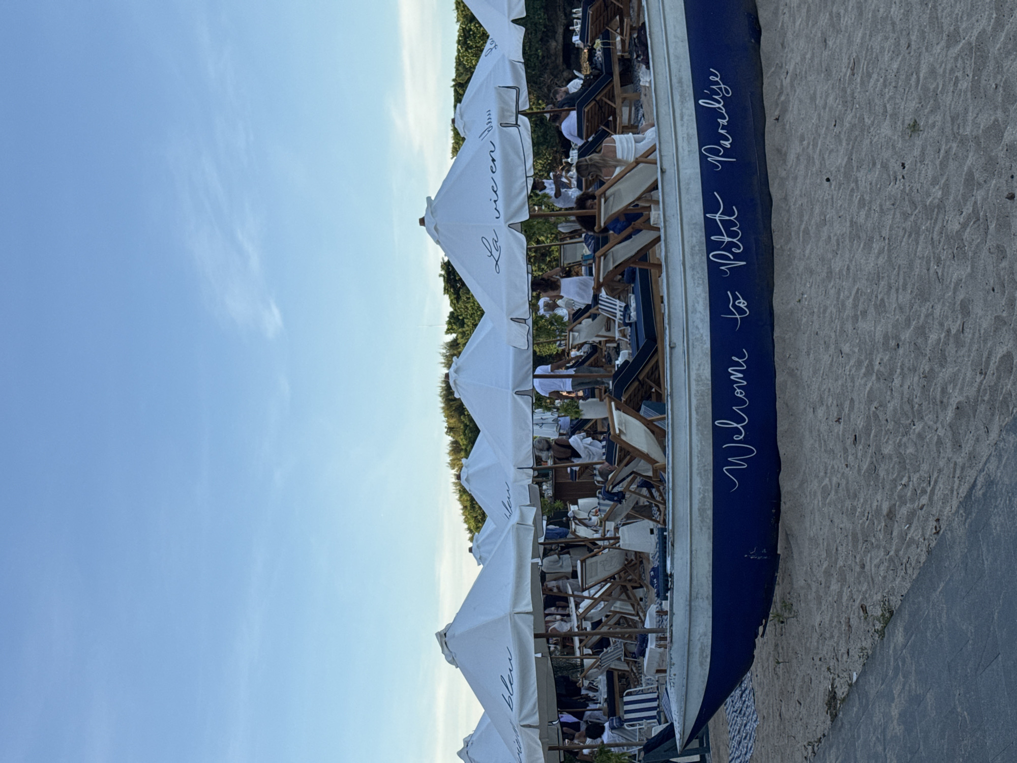 The image shows a beach scene, likely a private beach club in Kuta Selatan, Indonesia. A row of white beach umbrellas with text in French, such as "La Vie en Bleu", extends into the frame, shading numerous beach chairs. People are relaxing on the chairs. The scene has a clear blue sky. To the right is sand and a boat-shaped feature with "Welcome to Petit Paradise" written on it.