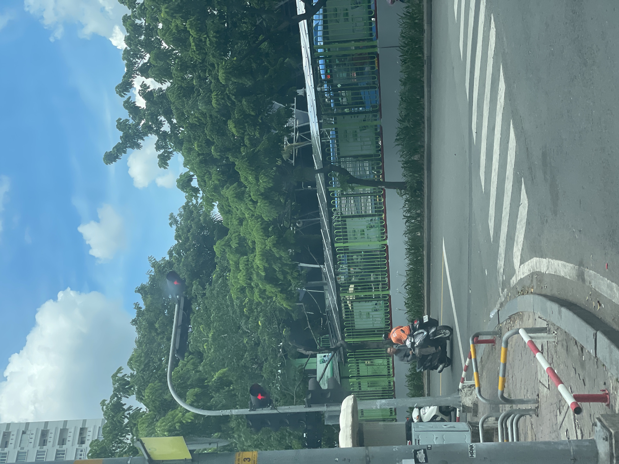 The image depicts a street scene in Thủ Đức City, Vietnam. Two individuals are on a black motorcycle, stopped at a red traffic light. The rider in front wears a dark jacket, and the passenger behind wears an orange shirt. A white scooter is parked to the left of the motorcycle. The street has a pedestrian crossing marked by white stripes. In the background, there are lush green trees, a building with multiple floors, a green fence with a patterned design, and a covered structure that appears to be a bus stop or shelter. The sky is blue with scattered white clouds. A traffic light signal is visible, displaying a red light. Street barriers with red, white, and yellow bands line the sidewalk.