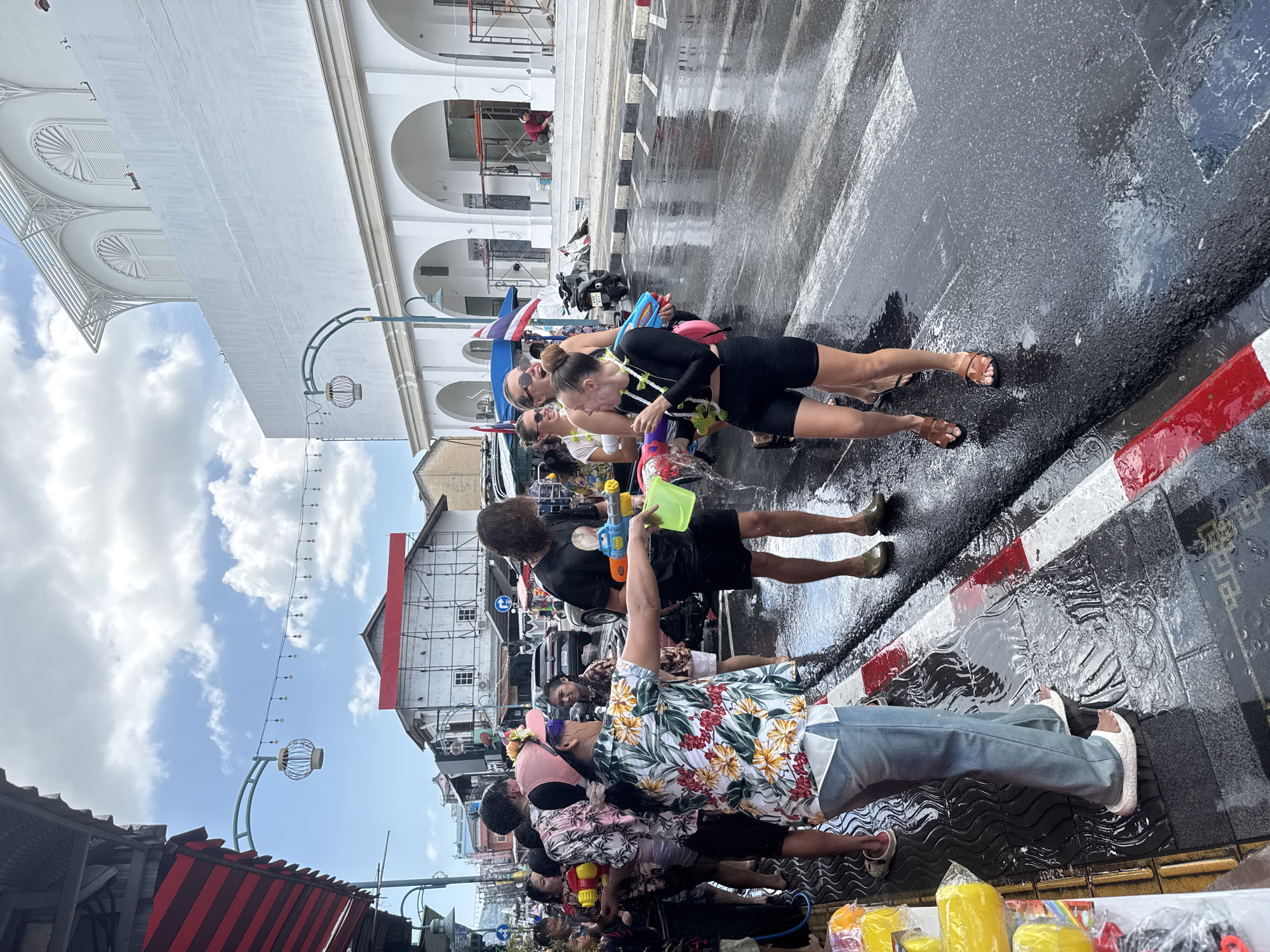 The image shows a street scene in Phuket City Municipality, Thailand. It appears to be during a water festival, as the street is wet and people are carrying water guns. A group of people are standing on a street corner near a curb. They appear to be soaking wet and holding water guns. The street is also wet, and there are puddles of water on the ground. One man is wearing a floral shirt and blue jeans. The buildings are mostly white with some decorative elements. The sky is blue with some clouds. There are also streetlights and wires visible. The pavement is wet.