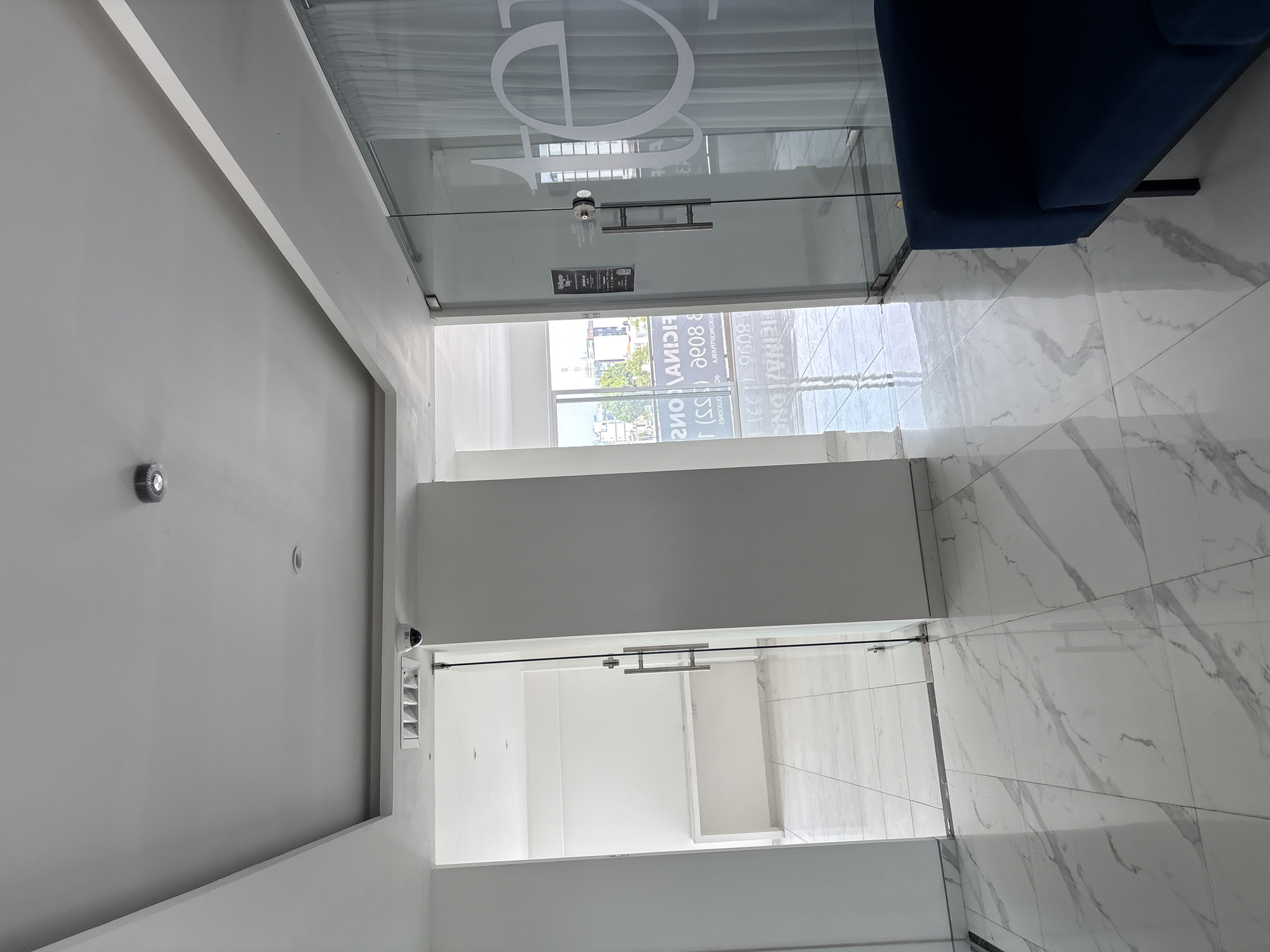 The image shows an interior space, likely a hallway or entrance area, in Puebla, Mexico. The area features white marble-like tile flooring with grey veining, suggesting a modern or upscale design. The walls and ceiling are white, contributing to a bright and clean aesthetic. There are two glass doors visible. The first door is a storefront-style glass door with a large, stylized logo, potentially "DO," printed in white. The second glass door is further down the hallway. The architecture is modern, with clean lines and an emphasis on light and open space. There is no clear indication of people or specific interactions in the scene. The environment is an indoor commercial or office space, likely a business establishment.
