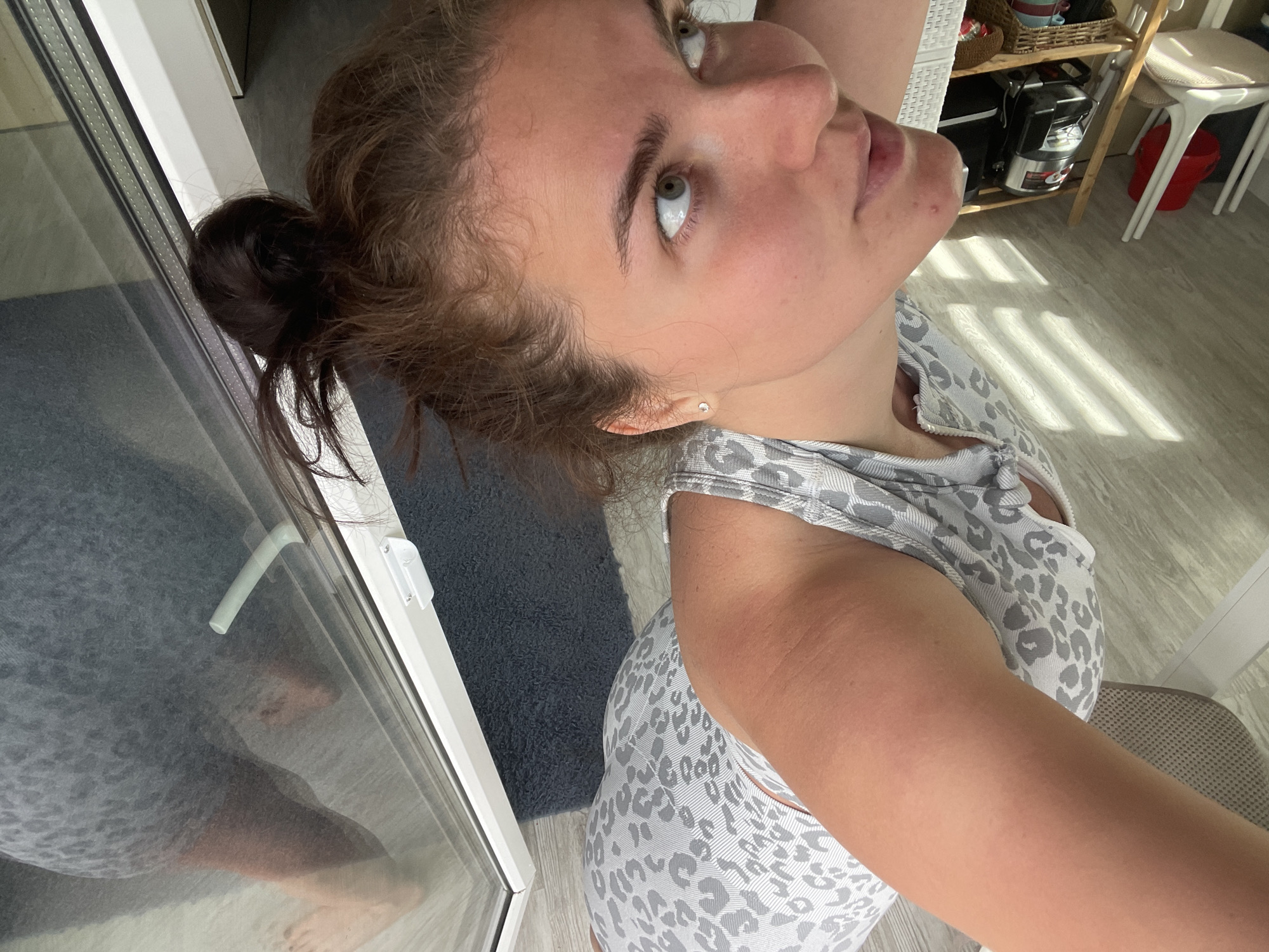A woman with brown hair in a bun is looking up towards the right of the frame. She is wearing a gray and white leopard print top. The background includes a door, a gray rug, light-colored flooring, a shelf with items on it, and a white chair. The image was not taken in the city of Мосметаллоконструкция Онт, Russia.