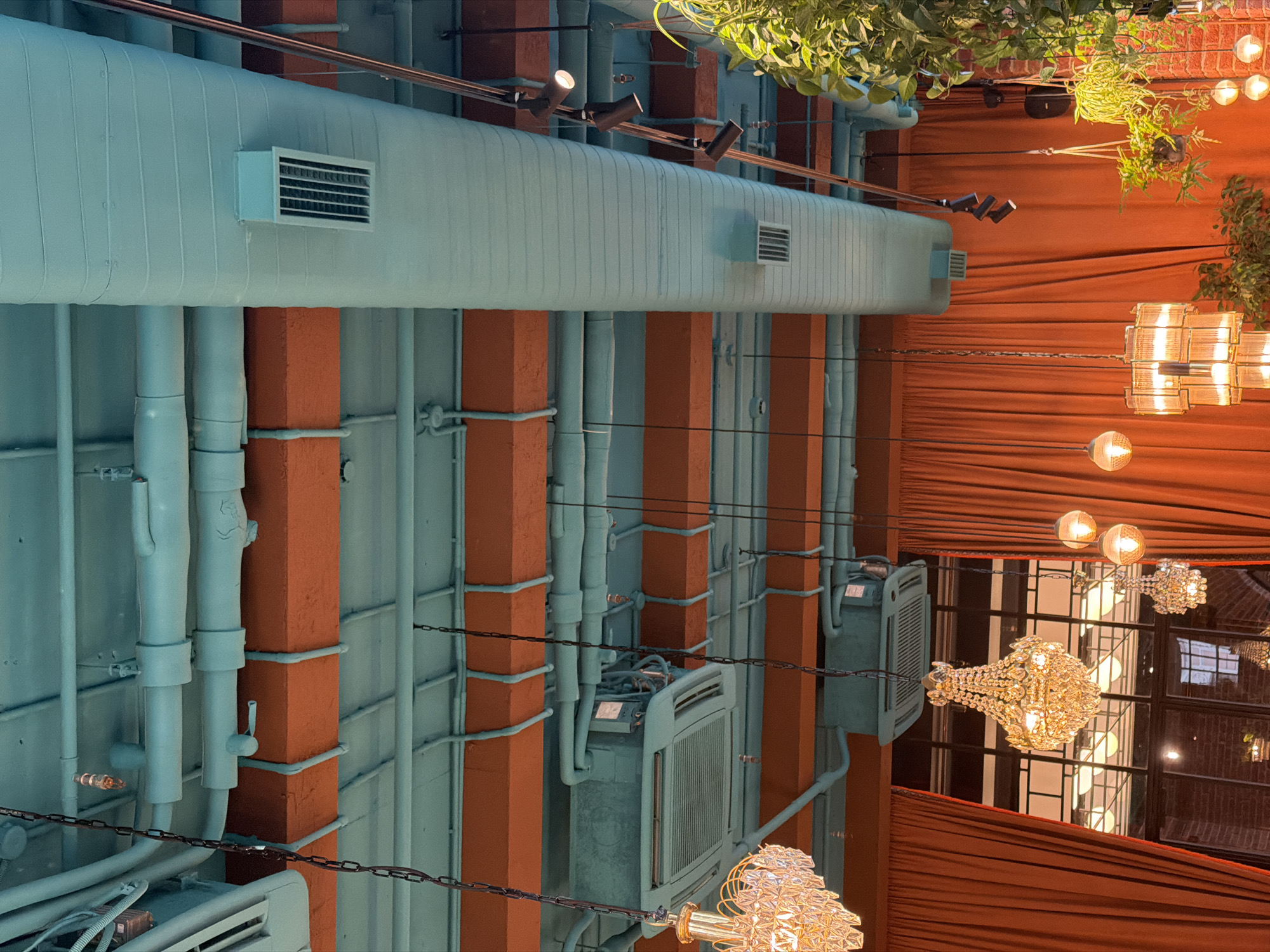 The image depicts an indoor scene, likely a restaurant or event space, with an industrial-chic aesthetic. The dominant features are large, light blue pipes running horizontally and vertically, contrasted with bright orange structural beams and dark orange drapery. Several air conditioning units are visible, mounted on the walls or ceilings. Hanging from above are decorative light fixtures, including ornate crystal chandeliers and simpler glass globe lights, suggesting an evening or dimly lit atmosphere. Green foliage is visible at the top of the image, cascading from an unseen source, adding a touch of nature to the industrial setting. The overall impression is one of a stylish, contemporary interior designed with a blend of raw and elegant elements. The orientation of the photo is vertical, with the ceiling appearing at the bottom and the floor at the top. No people are visible in the image.