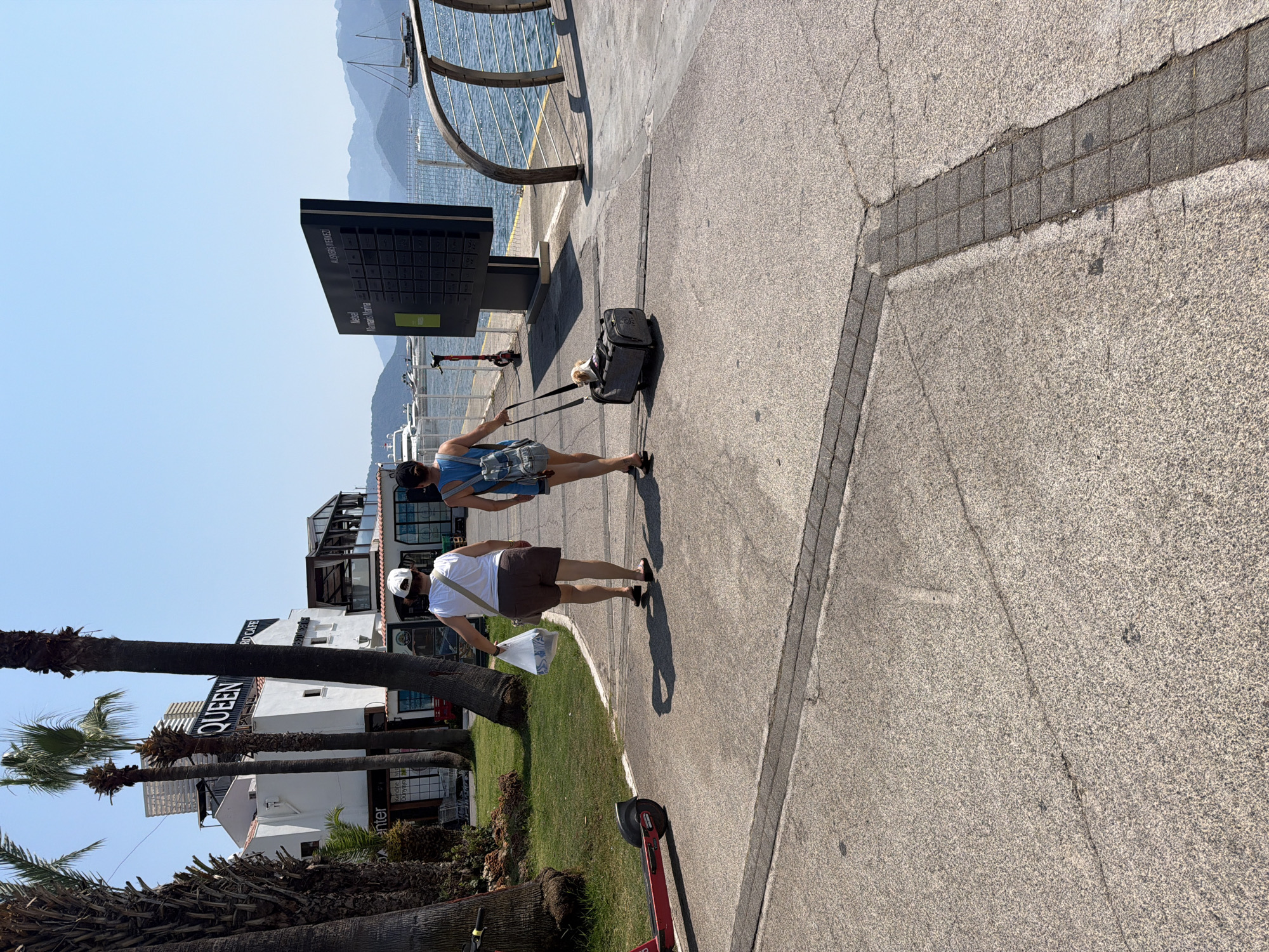 Two individuals are walking away from the camera on a paved walkway by the waterfront in Marmaris, Turkey. The person on the left is wearing a white tank top and brown shorts, carrying a white plastic bag in their right hand. The person on the right is wearing a blue sleeveless top and denim shorts, with a backpack and a wheeled pet carrier that contains a small dog. To the left of the walkway, there is a grassy area with palm trees and a white building with the word "QUEEN" prominently displayed. A red electric scooter is parked on the grass to the left. To the right of the walkway is a body of water with several boats and a large information board with Turkish text. The sky is clear and blue, and distant mountains are visible.