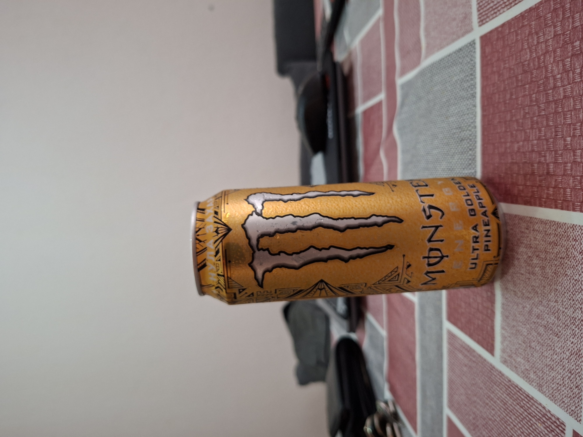A single Monster Energy drink can, oriented horizontally, is the main subject of this indoor photograph, taken in Plovdiv, Bulgaria. The can is a vibrant golden yellow with a textured finish, prominently featuring the iconic silver and black Monster "M" claw logo. Visible text on the can clearly states "MONSTER" and "ULTRA GOLDEN PINEAPPLE," with "ENERGY" also partially readable.
The can rests on a distinctive surface composed of a grid pattern of squares. These squares alternate between shades of red, grey, and white, with many exhibiting a finely woven or checkered texture, suggesting a tablecloth or a similar patterned surface. In the background, blurry dark shapes, possibly parts of furniture or equipment, are discernible against a plain, light grey wall. The lighting suggests an indoor setting, and no people are visible in the image. The can appears stationary, implying no specific activity is currently taking place.