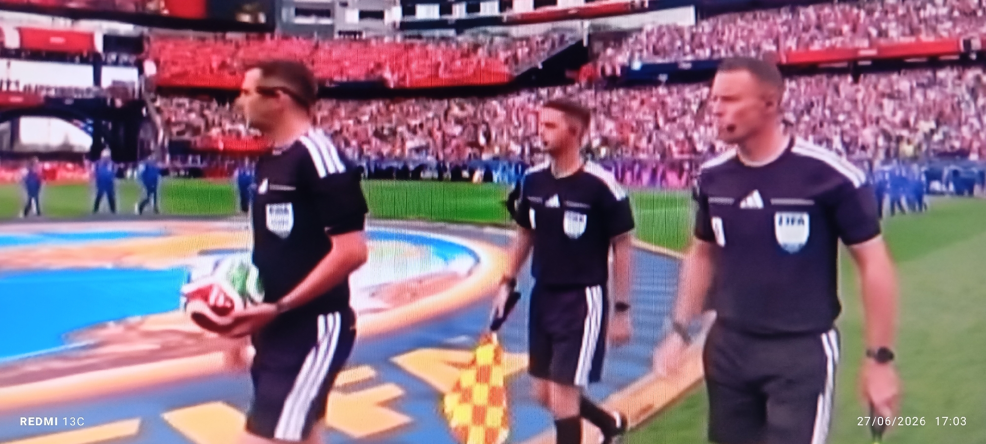 Three referees in black uniforms with white stripes are walking on a blue and yellow patterned surface, possibly a ceremonial pathway or field marking, in a stadium. The referee on the left is holding a soccer ball and appears to be entering the field. The referee in the middle is walking, and the referee on the right is also walking. Behind them, a crowd is visible in the red stands of the stadium. The image has a timestamp overlay of "27/06/2026 17:03" and "REDMI 13C" in the bottom left corner. The scene depicts a sporting event with referees preparing to officiate. There is no information available to determine the location in the city of Idimu, Nigeria, as this is a generic stadium environment.