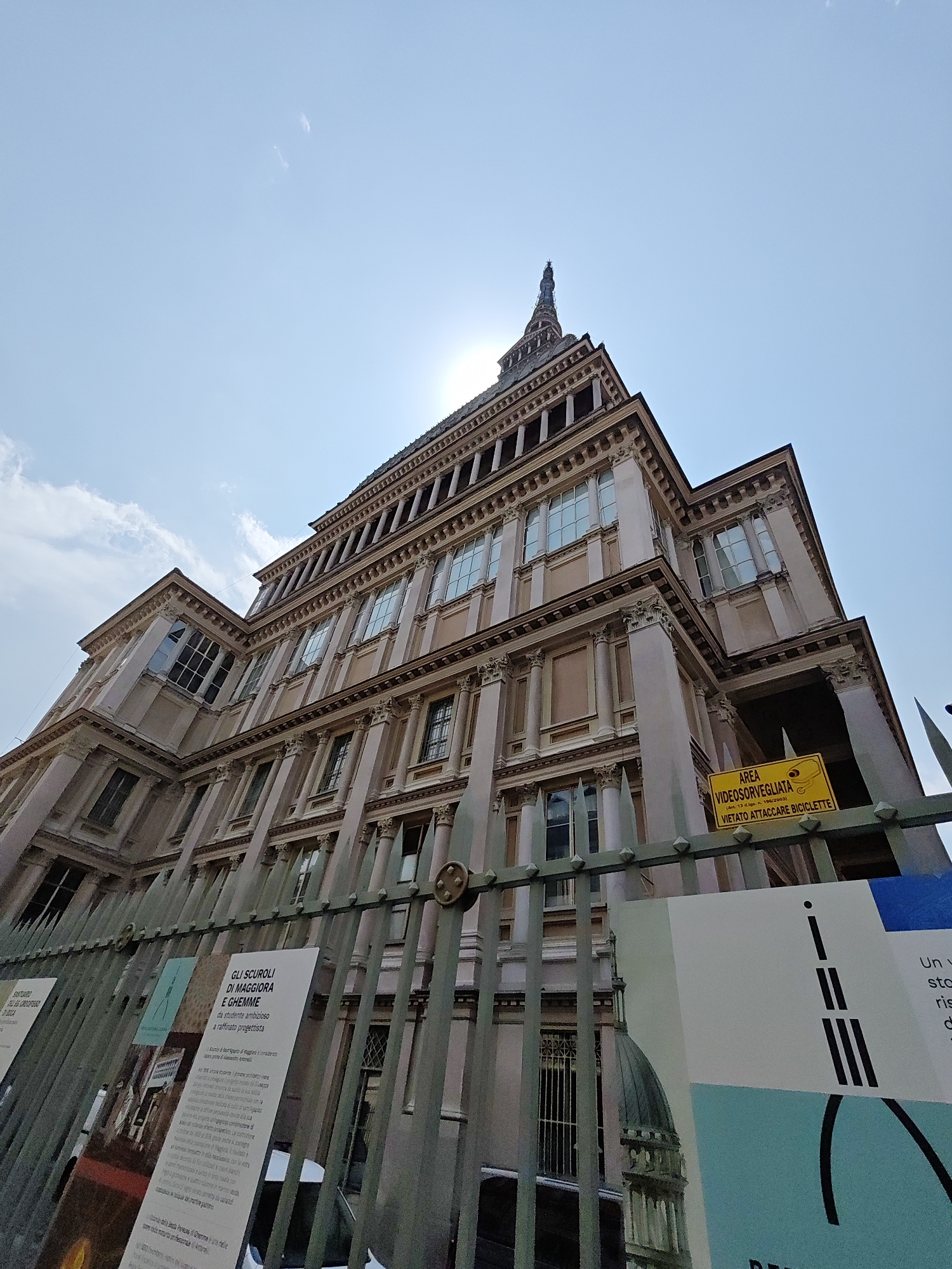 The image shows the Mole Antonelliana in Turin, Italy, viewed from a low angle. The sun is shining brightly, with some clouds in the sky. The Mole Antonelliana is a prominent landmark in Turin, known for its distinctive spire. The building is made of stone and has many windows. In the foreground, there is a metal fence with several signs attached to it. One sign is yellow and reads "AREA VIDEOSORVEGLIATA VIETATO ATTACCARE BICICLETTE," which translates to "AREA UNDER VIDEO SURVEILLANCE. BICYCLES PROHIBITED." Another sign is white and has Italian text and images related to an exhibition. The scene appears to be a public space, possibly a plaza or street in front of the building.