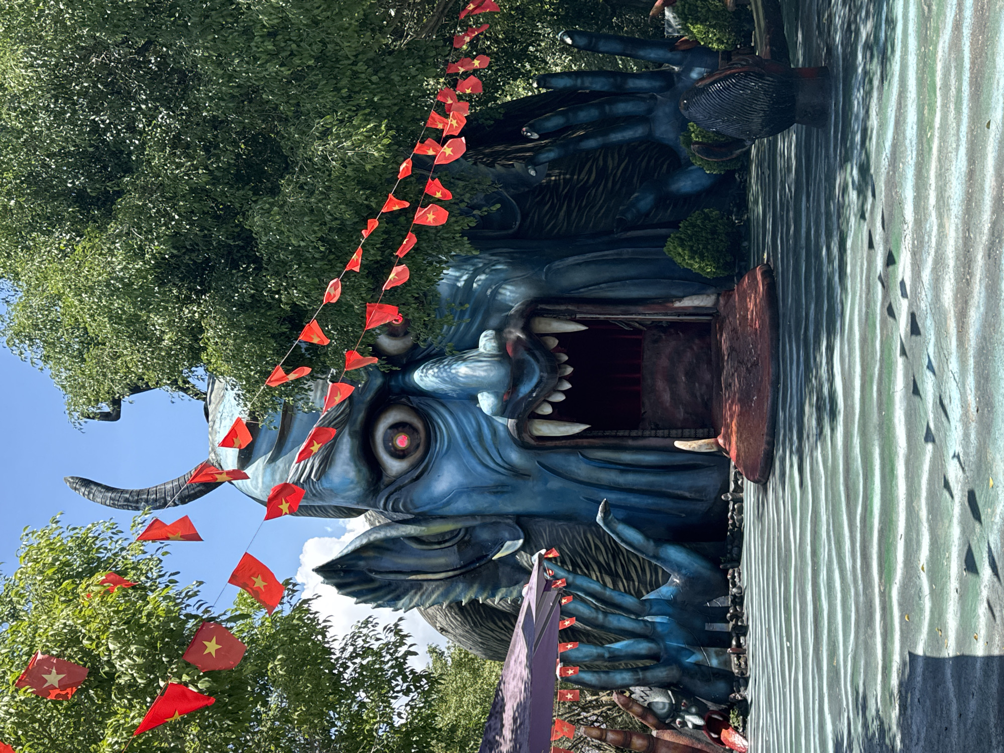 The image shows a large, blue, monstrous sculpture with sharp teeth and a gaping mouth, set against a backdrop of lush green trees and a clear blue sky. Red and yellow triangular flags, likely Vietnamese flags, are strung across the scene, adding a festive element. The sculpture's hands are large and blue, with visible fingers. In front of the sculpture is a paved area designed to resemble water with rippling patterns. There appear to be other smaller sculptures in the foreground. The overall scene suggests an outdoor attraction or theme park, possibly located in Păng Tiêng 1, Vietnam, given the presence of the Vietnamese flags. No people are visible in the image.