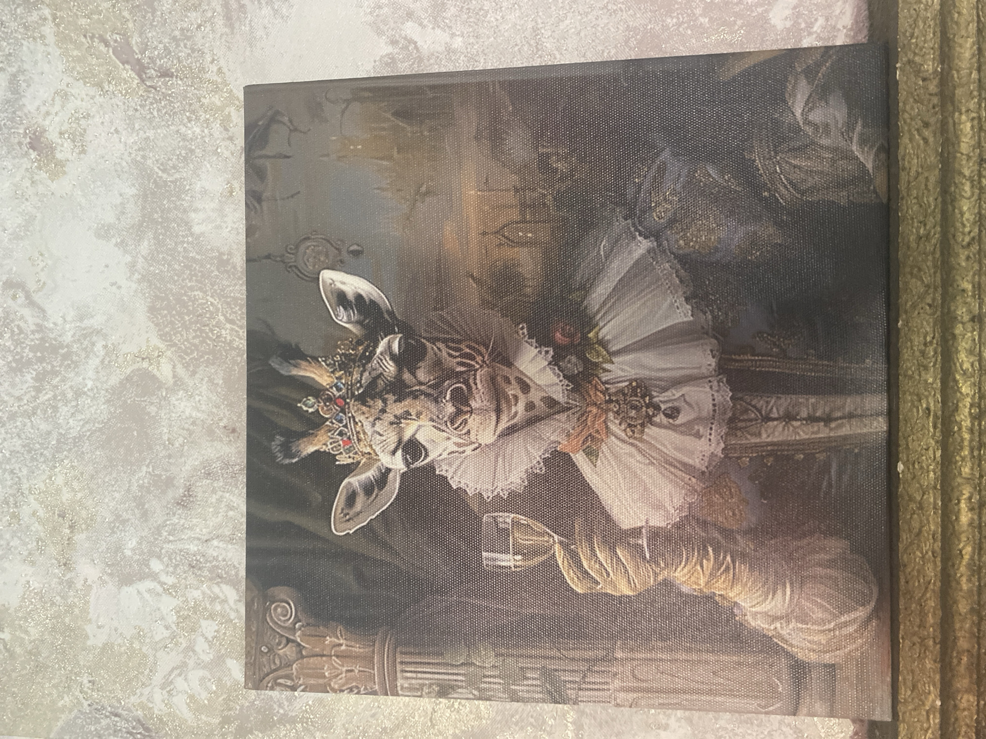 The image is a close-up, overhead shot of a canvas print of a painting. The painting depicts a giraffe wearing a crown and ornate royal attire, holding a glass of white wine. The giraffe has a regal expression and is adorned with a ruffled collar, jewels, and a sash. The background of the painting features architectural elements that suggest a castle or palace, with a soft, hazy atmosphere. The canvas print is framed by a decorative, golden border. The background behind the canvas appears to be a textured wall with a light, speckled pattern. The exact location in Blagoevgrad, Bulgaria, is not discernible from the image.