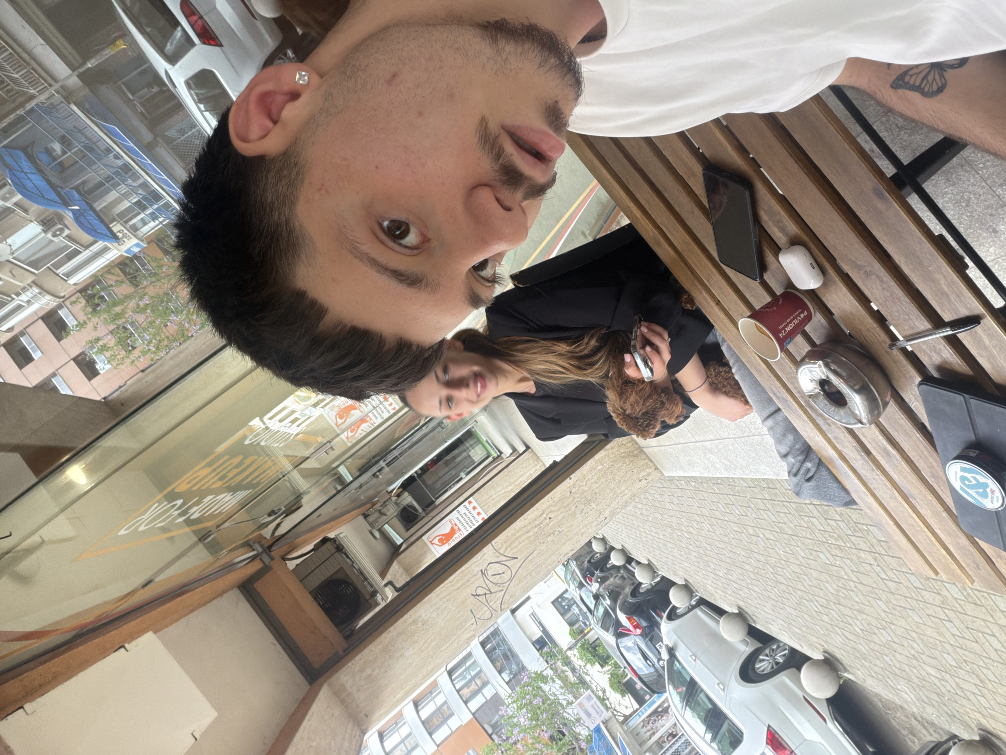 The image shows a man and a woman seated at a wooden table, likely at an outdoor cafe. The man, who appears to be taking the picture, has short dark hair, a goatee, and is wearing a white shirt. The woman has long brown hair and is holding a small brown dog. On the table, there is a mobile phone, Airpods, a cup of coffee, an ashtray, a pen, and a weighing scale. In the background, there is a cafe window and a street with buildings and cars. Based on the objects on the table and the architectural style of the building in the background, it is likely that the location is in a city and based on the PAVILION24 cofee cup I assume that location is in the city of Sofia, Bulgaria.